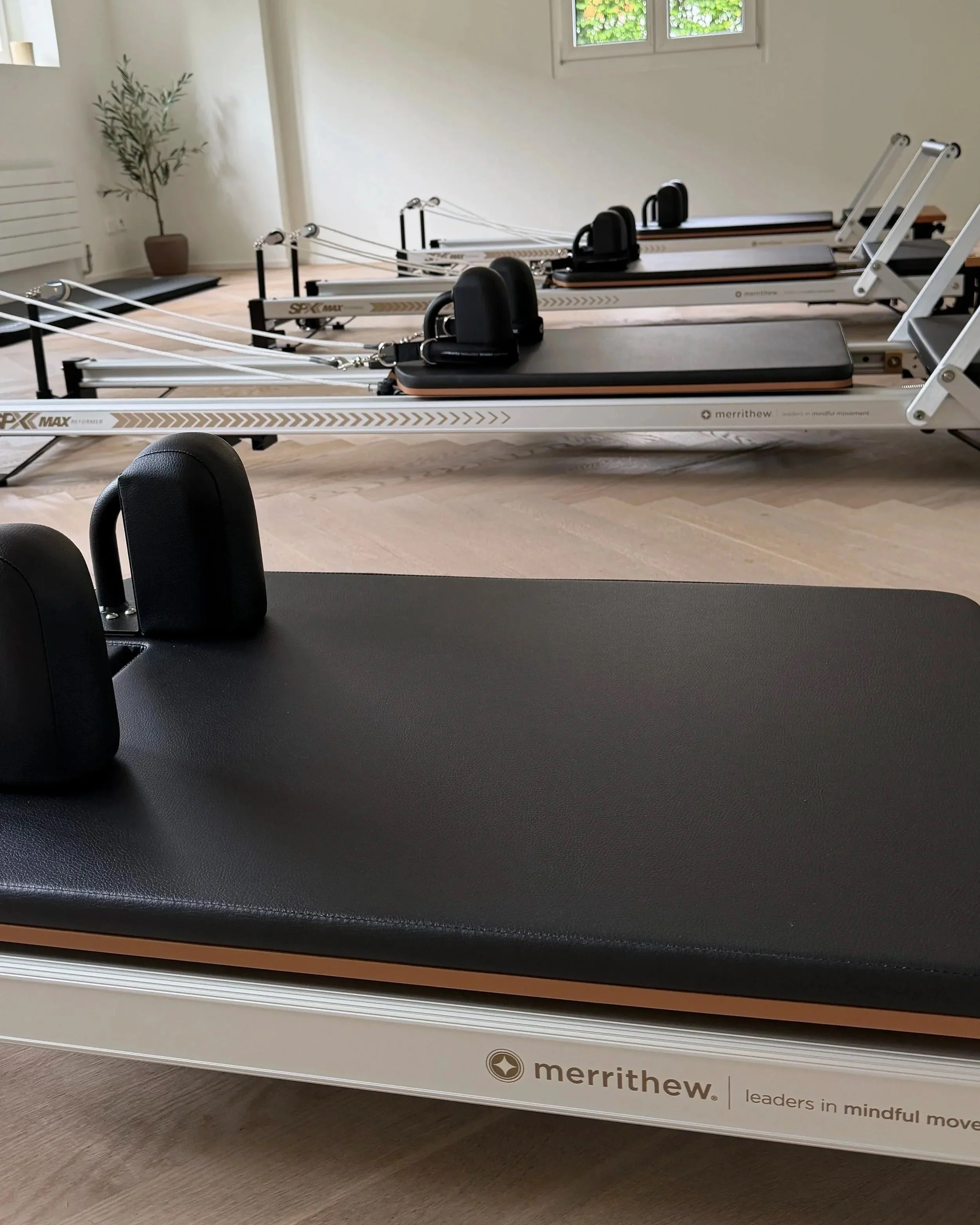 Mehrere Pilates-Reformer in einem Raum, das Fenster im Hintergrund zeigt Grünpflanzen.  'Merrithew' Refornerund mit Slogan 'leaders in mindful movement'.