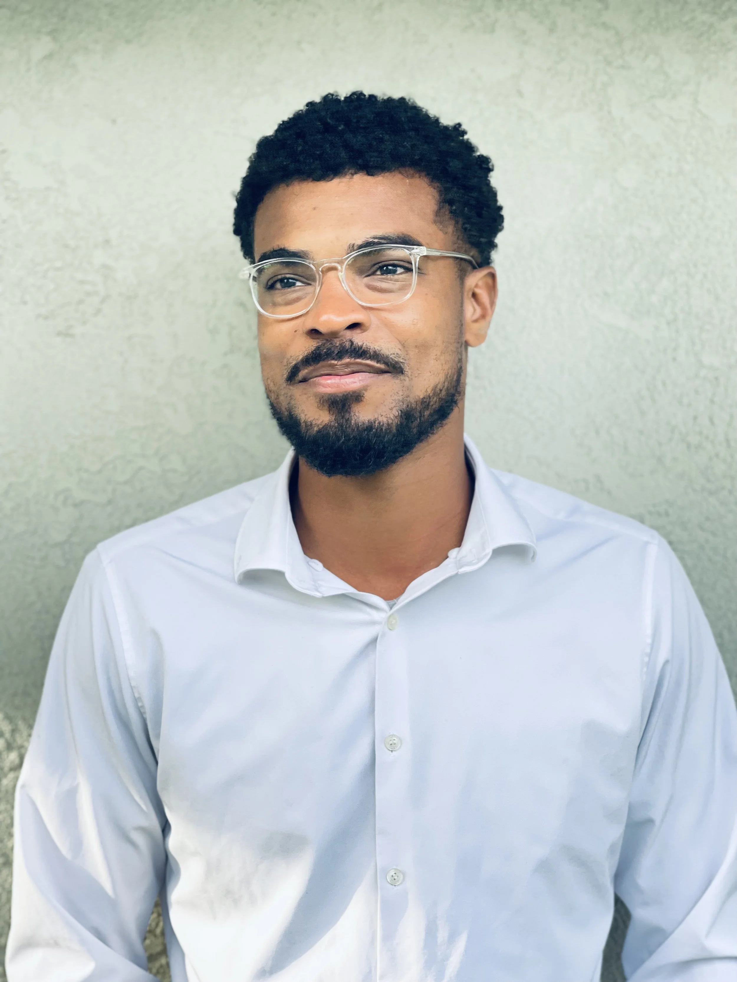 A man with glasses and a beard wearing a white dress shirt standing against a textured light-colored wall.