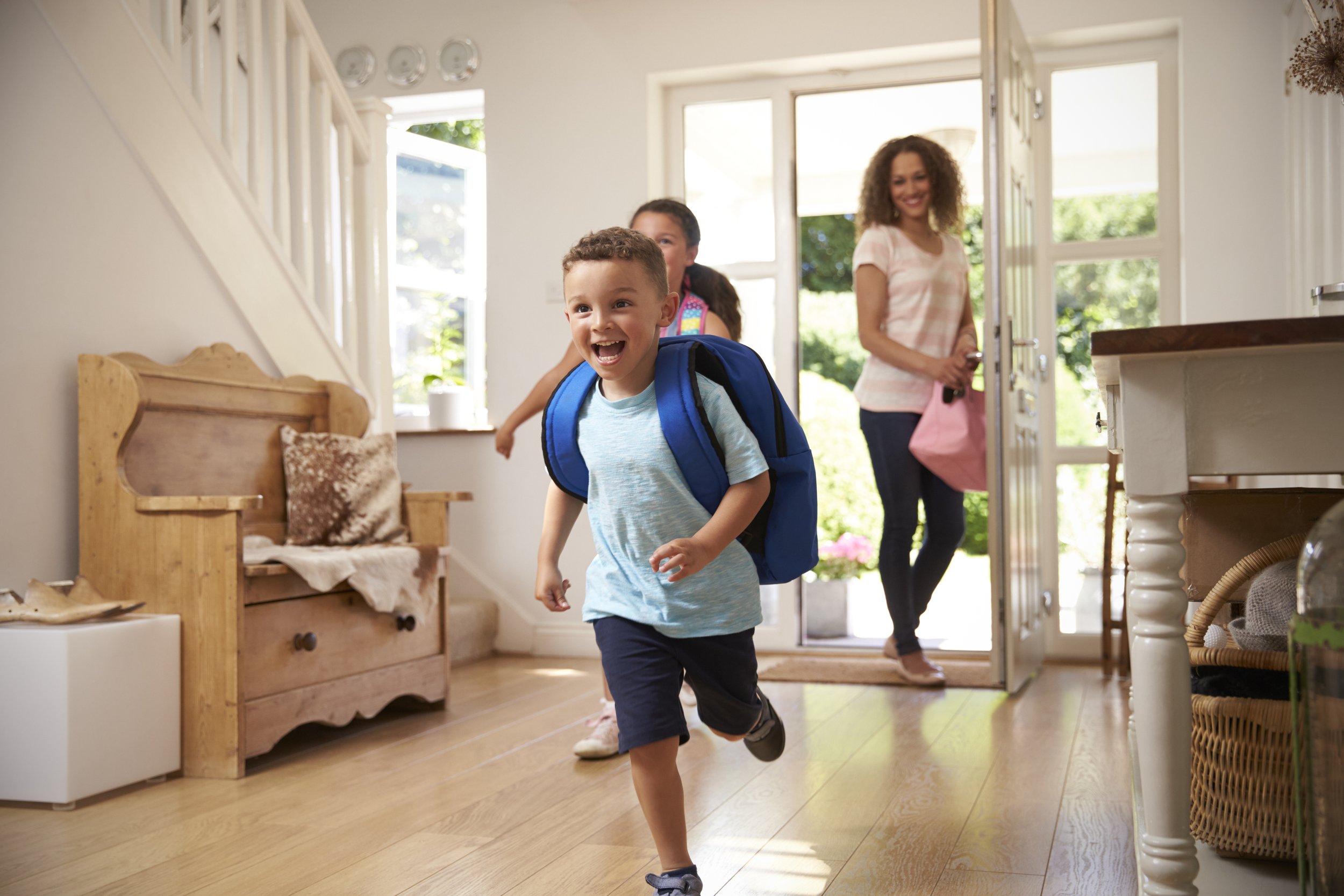 A young boy with a blue backpack running inside a bright house, followed by a woman and a girl, near the front door.