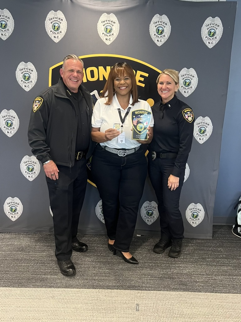 Three police officers, including two women and one man, standing together, smiling, during an award or recognition ceremony at the Monroe Police Department, with a backdrop featuring Monroe Police badge logos.