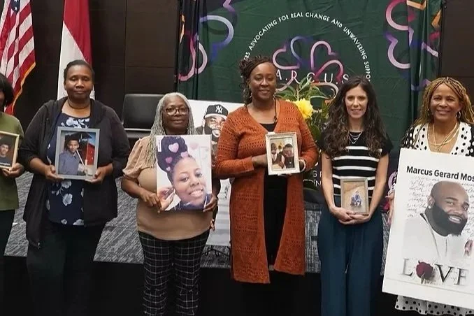 Group of five women standing indoors, each holding framed photographs. Behind them are banners and flags, including the American flag and a green banner with purple and pink graphics and text.