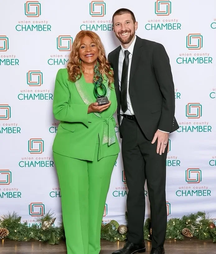 A woman in a green suit holding an award standing next to a man in a black suit, in front of a backdrop with the Union County Chamber logo and holiday decorations at the bottom.