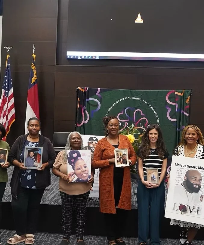 Group of six women standing in front of a banner at an indoor event, holding framed photographs and posters, with flags in the background.