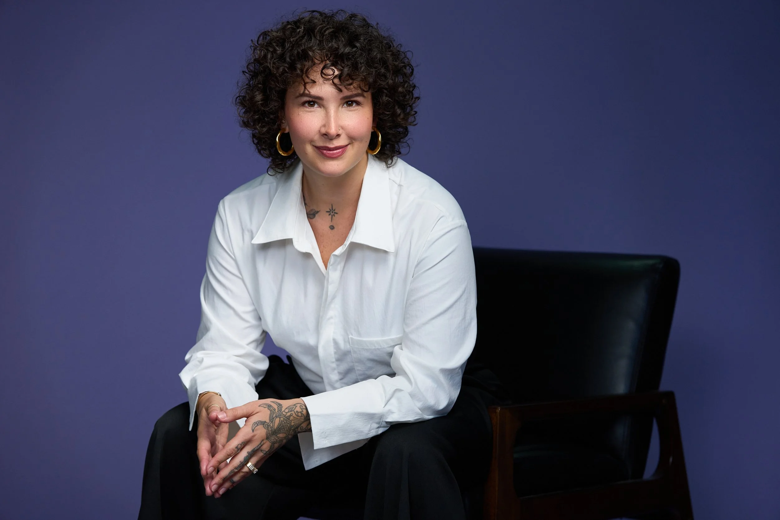A woman with curly dark hair, wearing gold hoop earrings and a white button-up shirt, sitting on a black chair against a purple background.