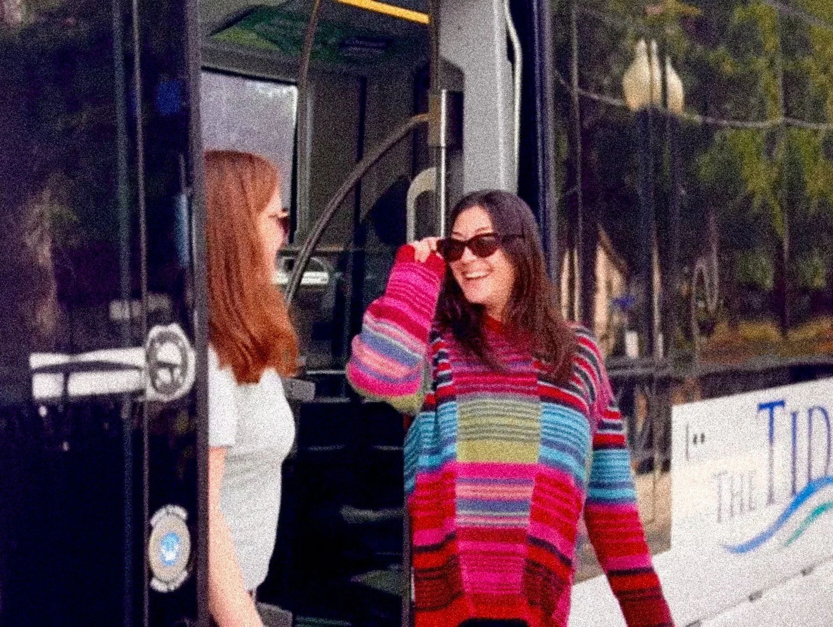 Two women standing outside a Tide Light Rail Vehicle, one with red hair wearing sunglasses and a white dress, the other with dark hair wearing a colorful striped sweater and sunglasses, smiling at each other.