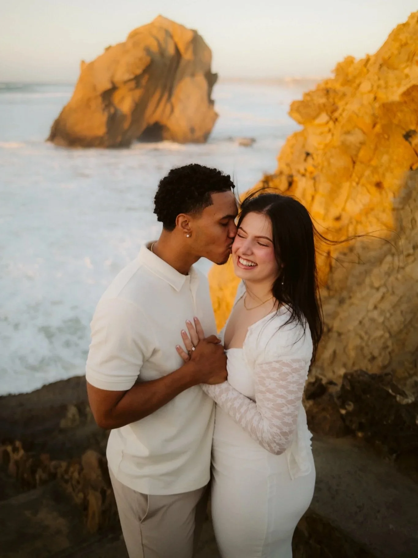 Couple playing on the beach with rocks in the background, she smiling, he kissing her on the forehead, at sunset.