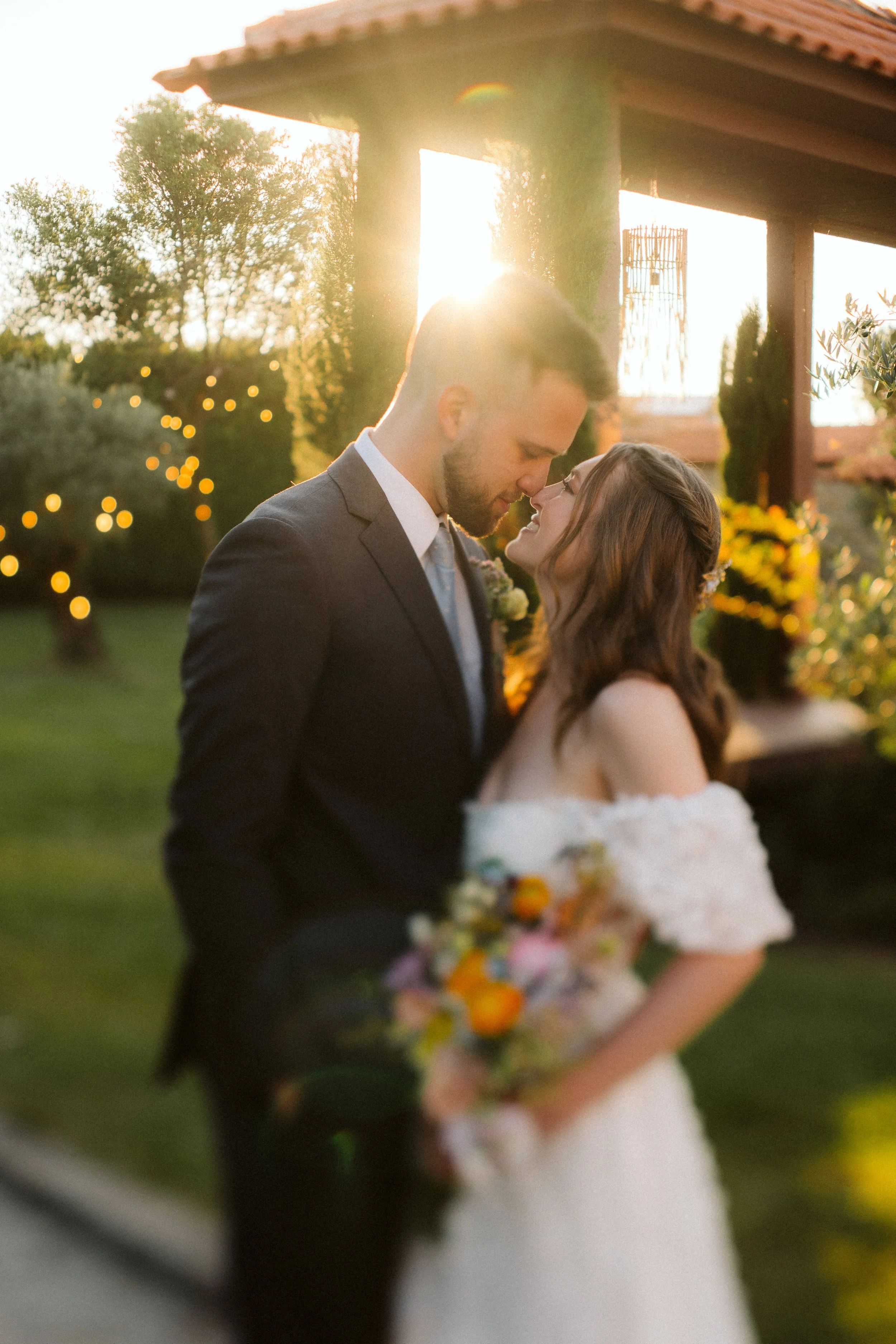 Couple standing smiling, about to kiss, at sunset with warm lighting, during an outdoor wedding ceremony.