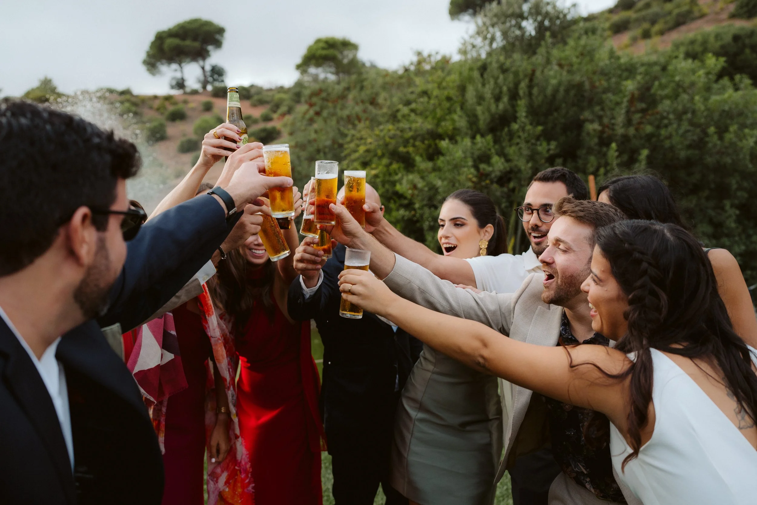 Group of people toasting with beer glasses outdoors in a natural setting, smiling and celebrating.