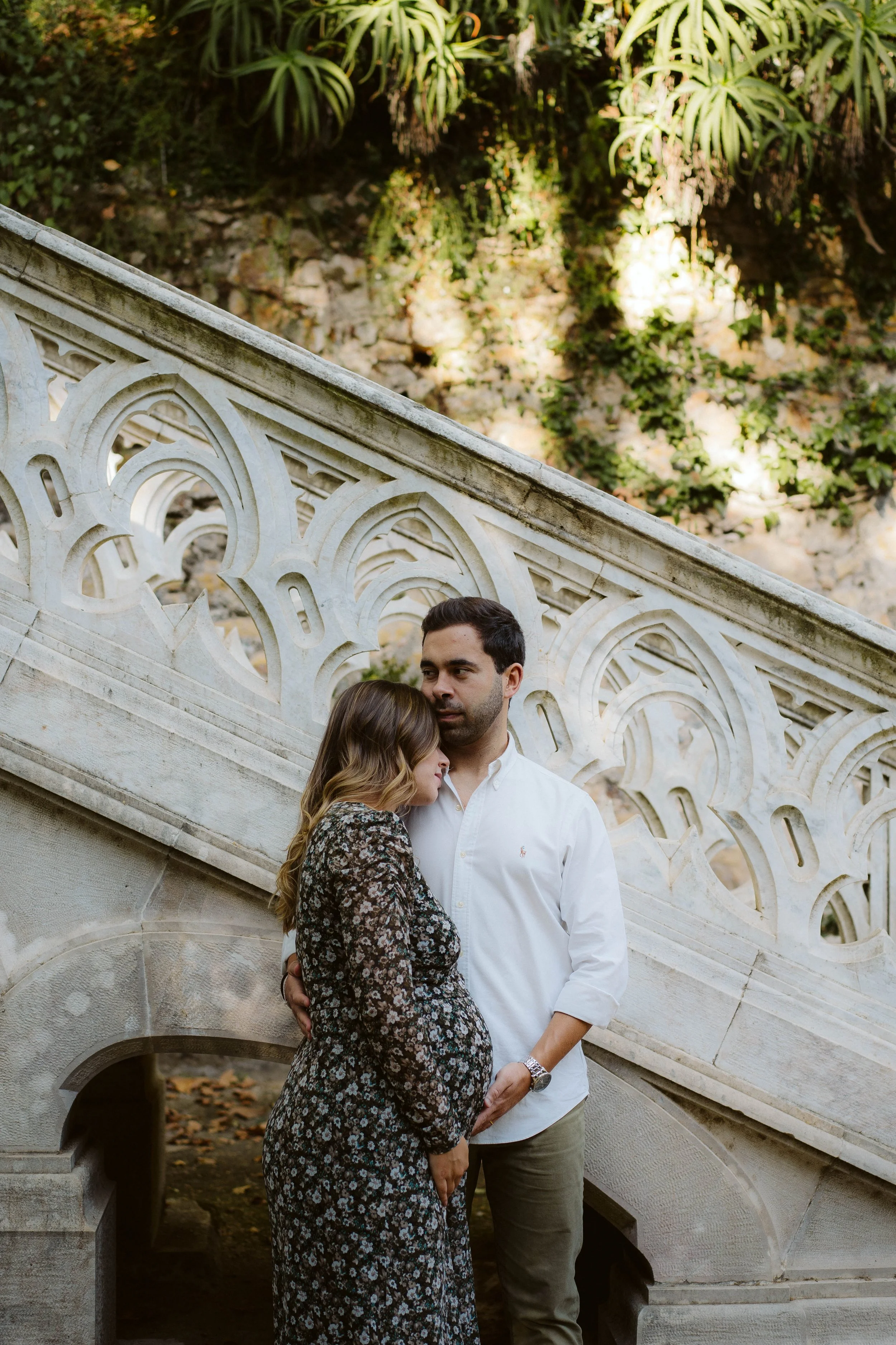 Pregnant couple embracing under a stone staircase in a garden with vegetation.