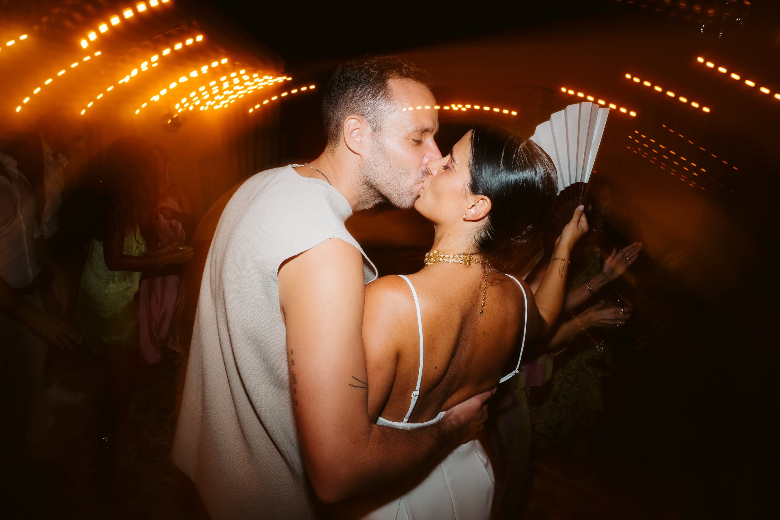 Couple passionately kissing at a wedding party, with warm yellow lighting and people in the background.