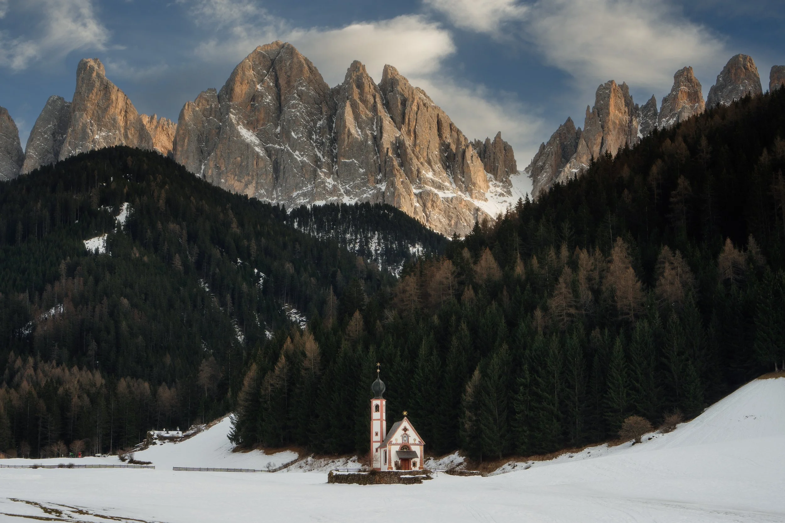 Scene of rocky mountains in the background, forest trees on the slope, and a small white chapel in the snow in the foreground, in Italy
