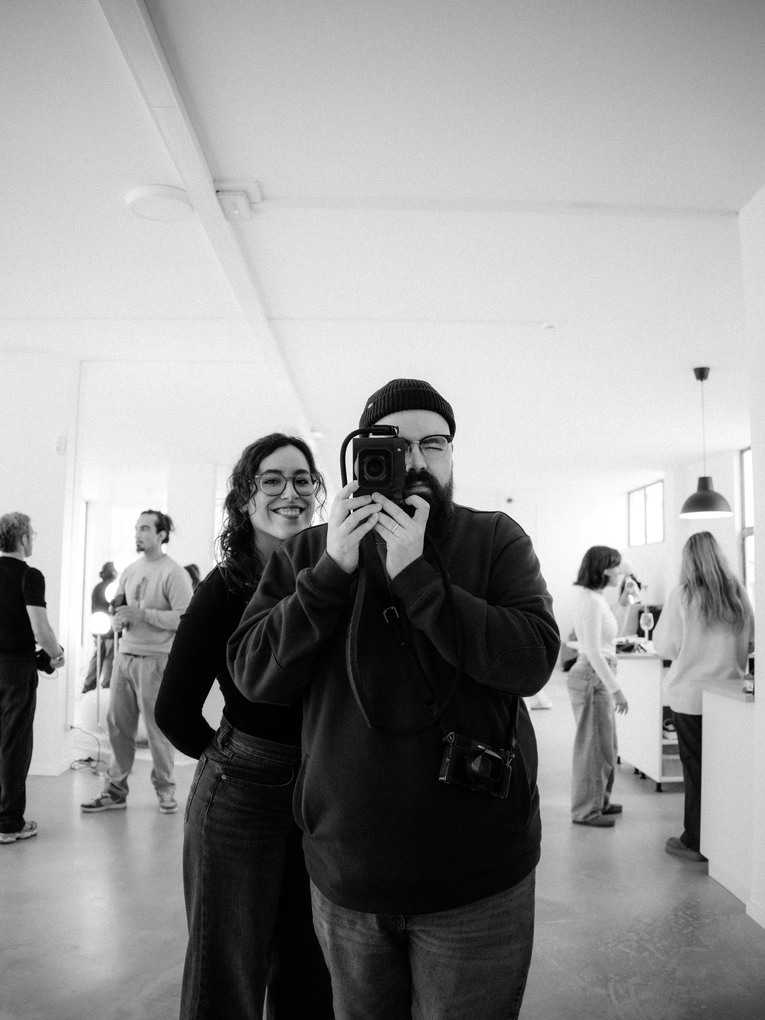 Black-and-white image of two people taking a selfie. The person in front, with a beard, glasses, and a beanie, holds the camera. The person beside them, with curly hair and glasses, smiles at the camera.