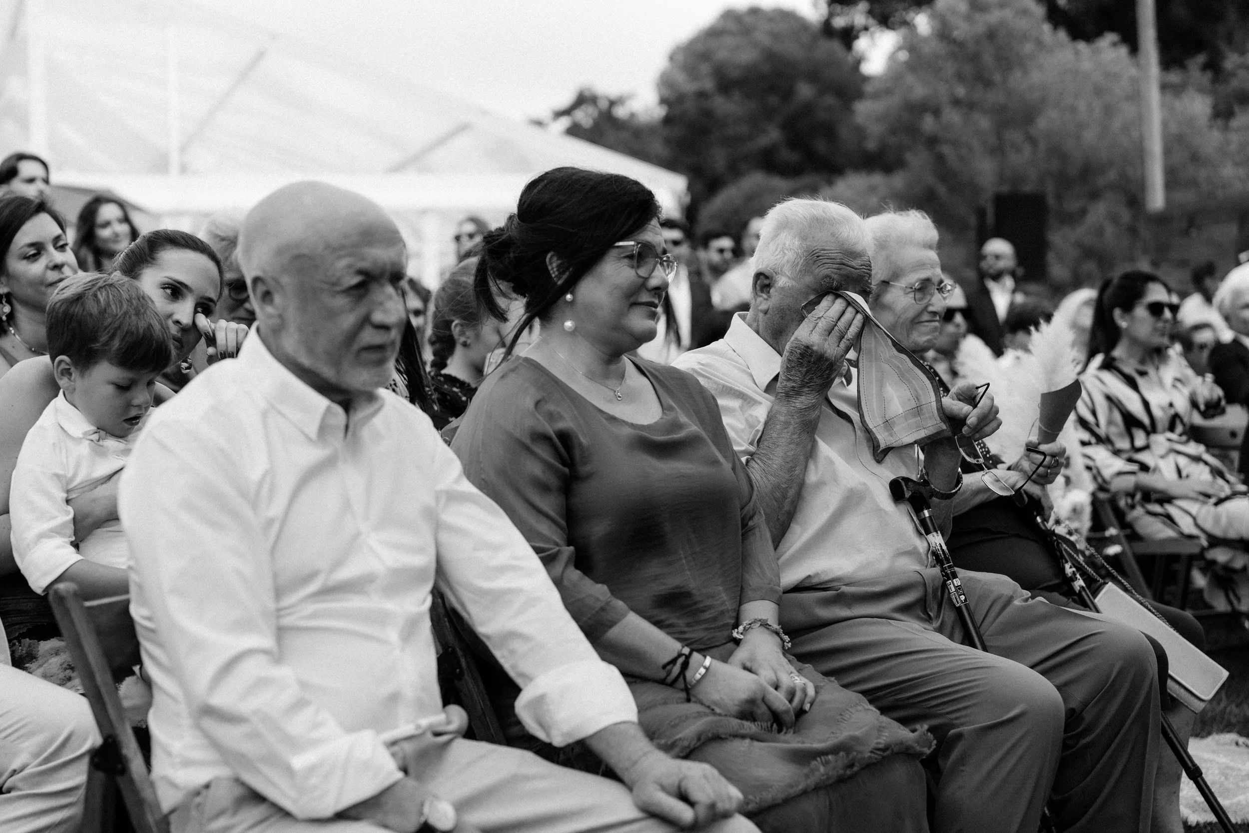 Group of people at an outdoor ceremony, including elderly, young adults, women, and children, seated in chairs, experiencing emotional moments, with one person covering their face with a handkerchief, in black and white.