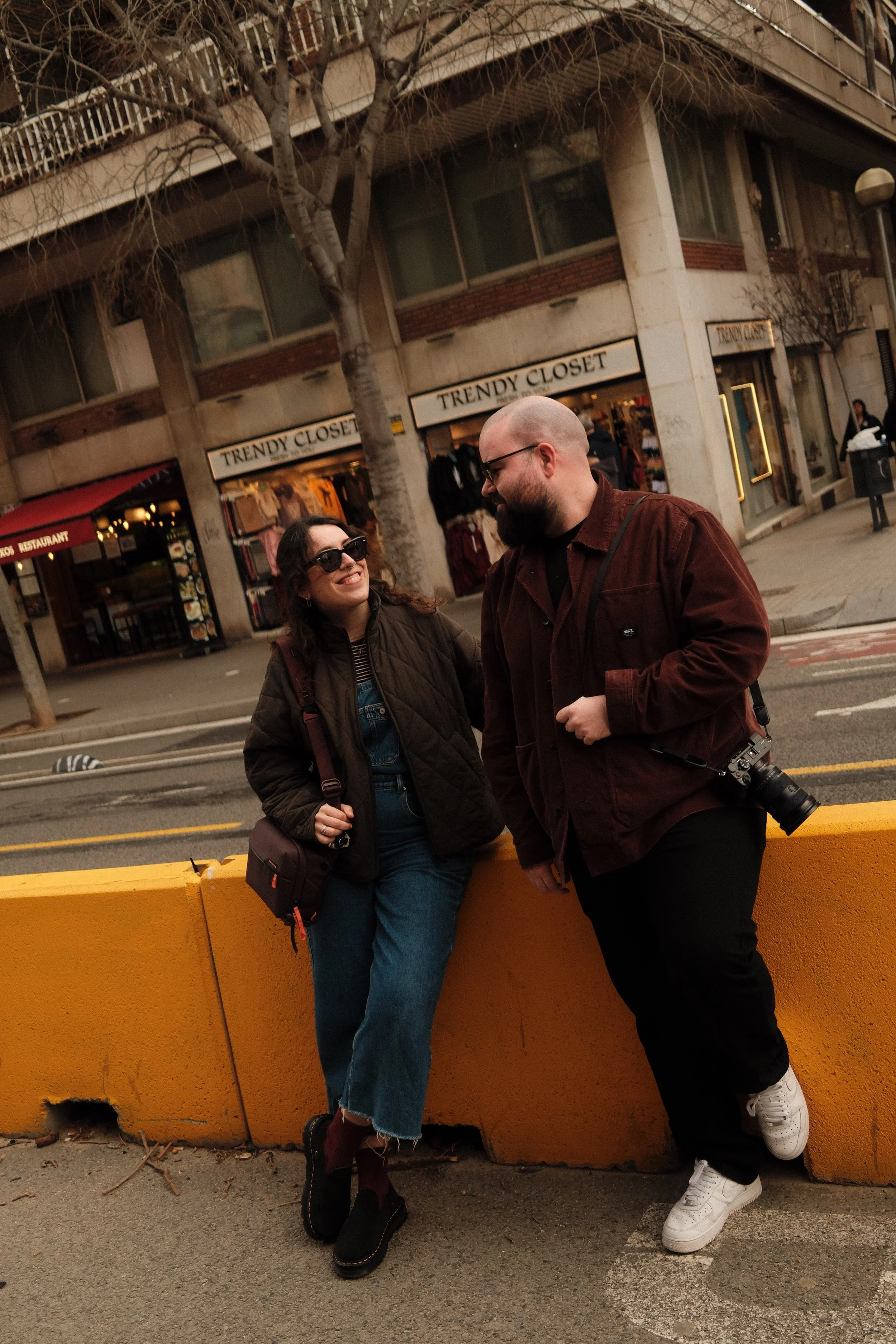 Man and woman talking and laughing on the streets of Barcelona, with clothing stores in the background, during the day.