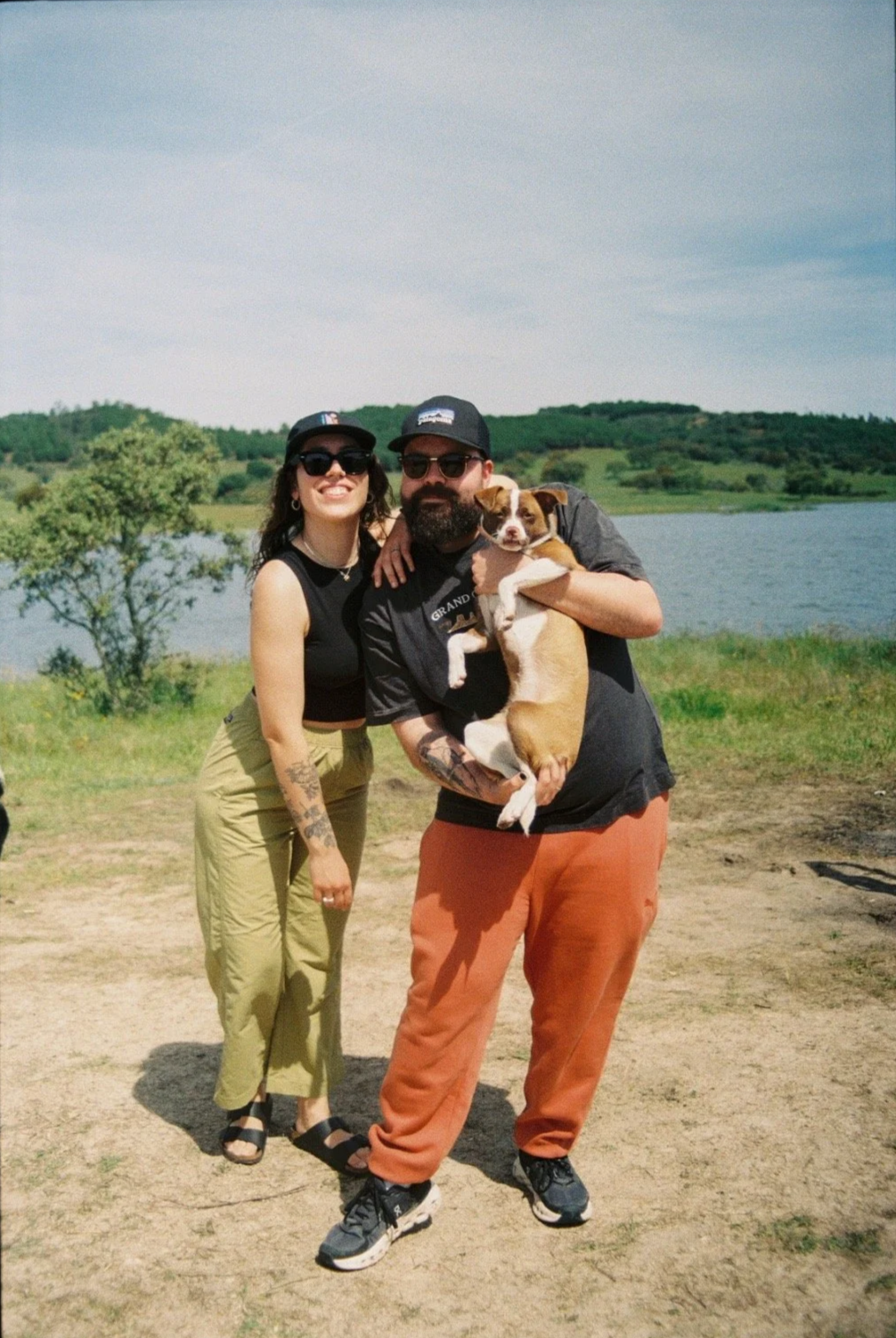 Smiling couple holding a dog near a pond, with vegetation in the background.