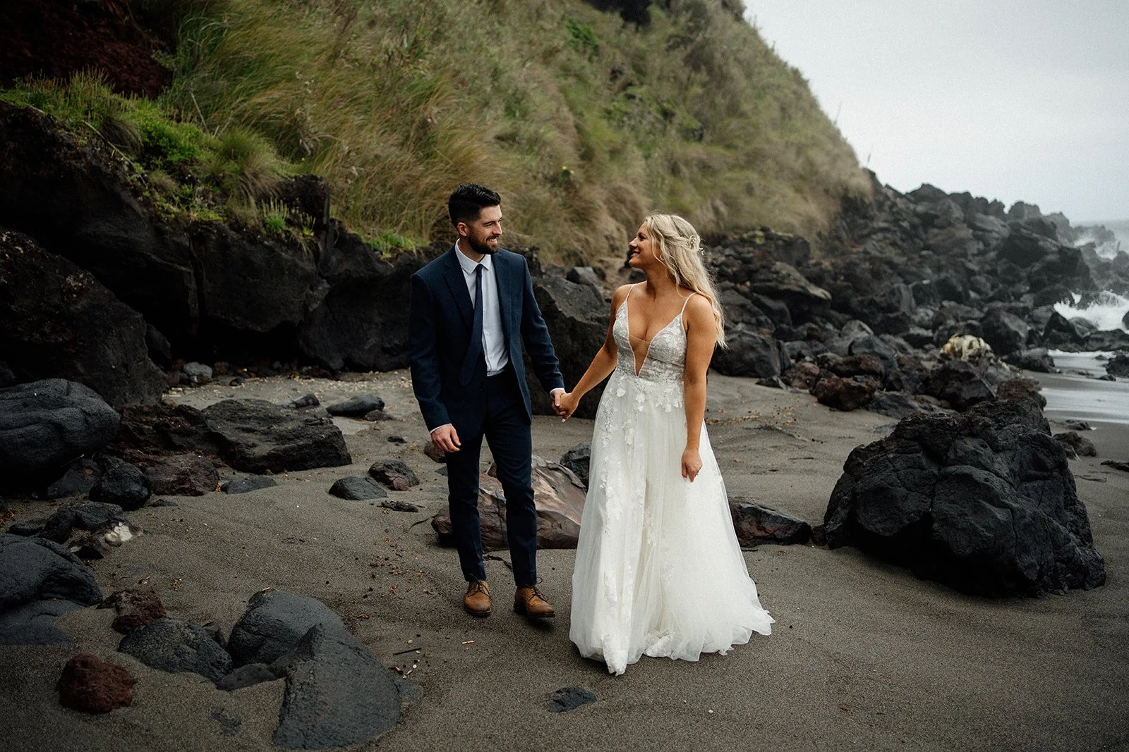 Wedding couple on the beach, walking hand in hand with cliffs and the sea in the background.