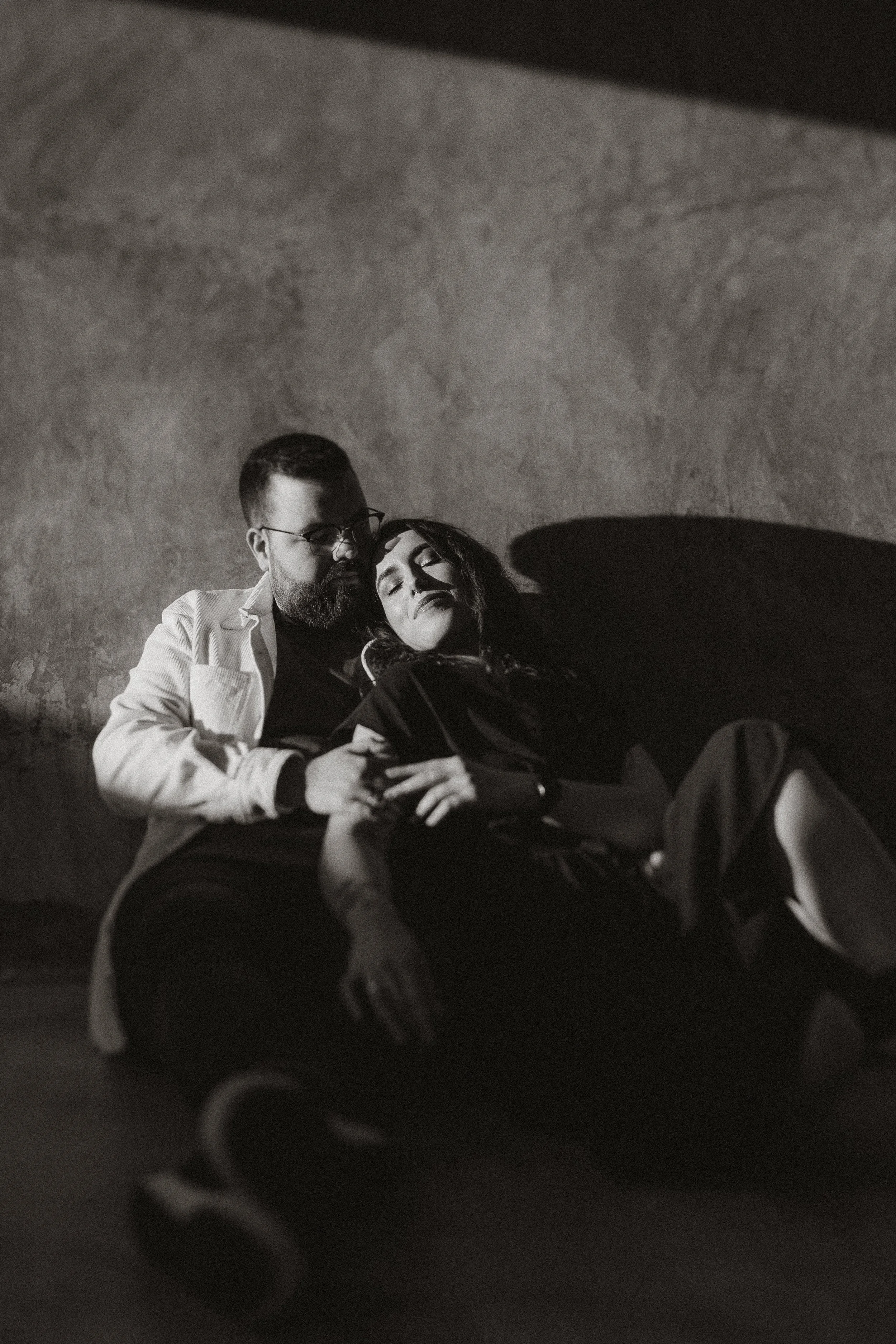 Man with sunglasses and a beard holding a woman with dark hair and a serene expression, both sitting on the floor, with a wall in the background.