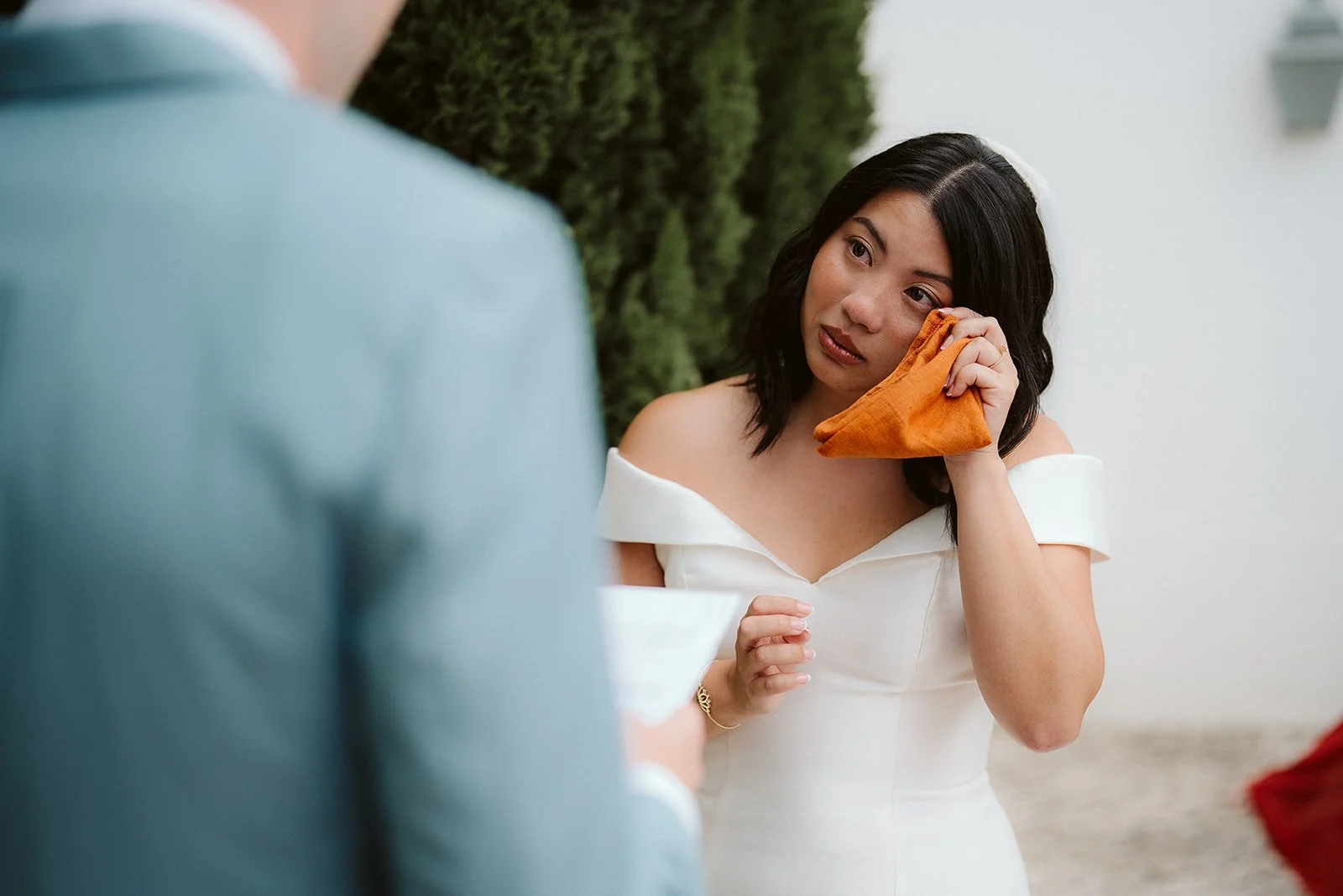 Woman in a white wedding dress with an emotional expression, holding an orange fabric near her face, in an outdoor setting.