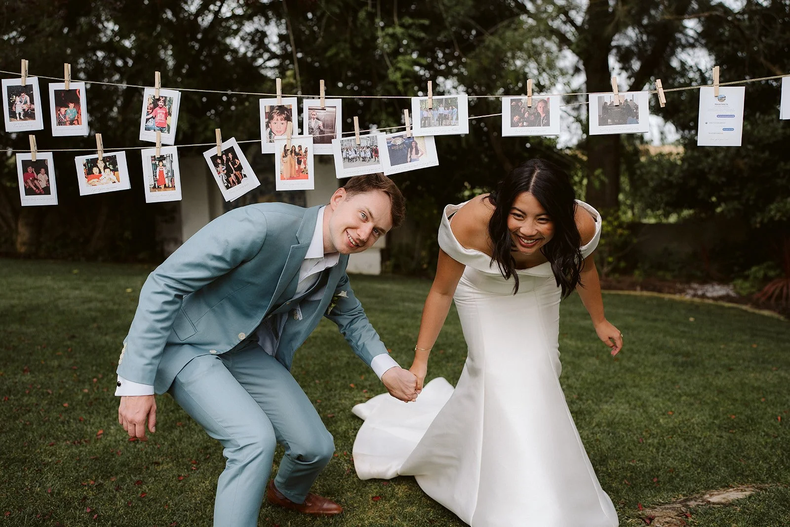 Wedding couple playing in the garden, holding hands and smiling, with photos of special moments hanging on a string in the background.