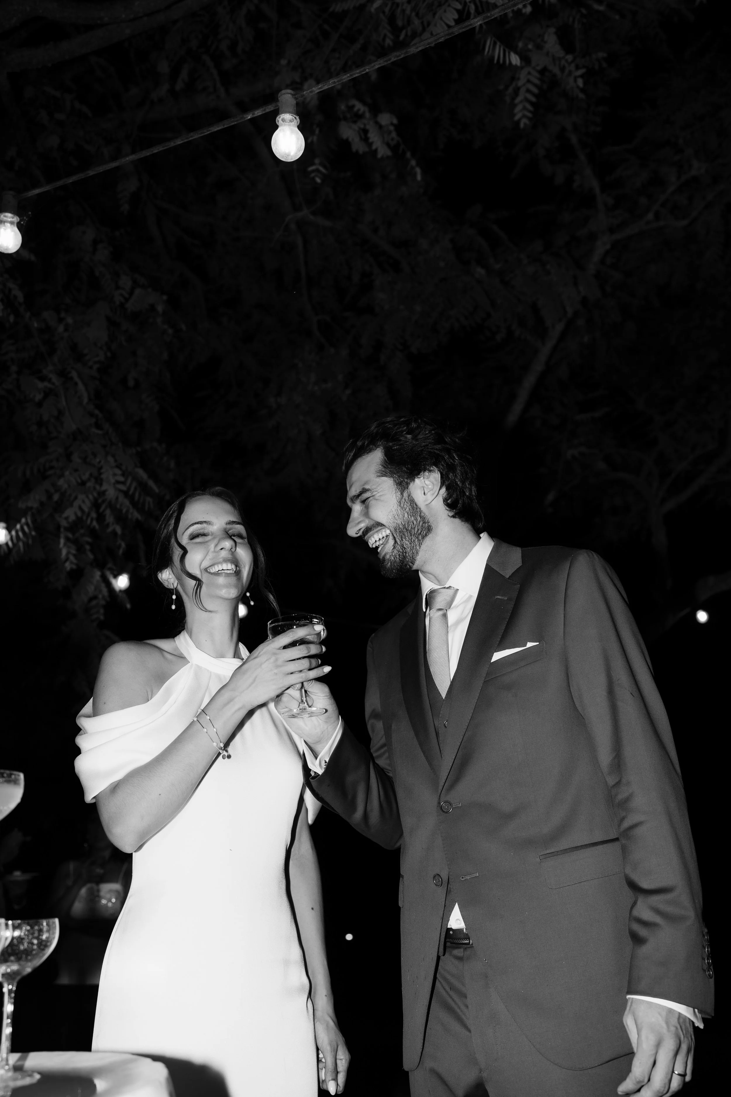 Smiling couple at a wedding, toasting with glasses of drinks, outdoors at night.