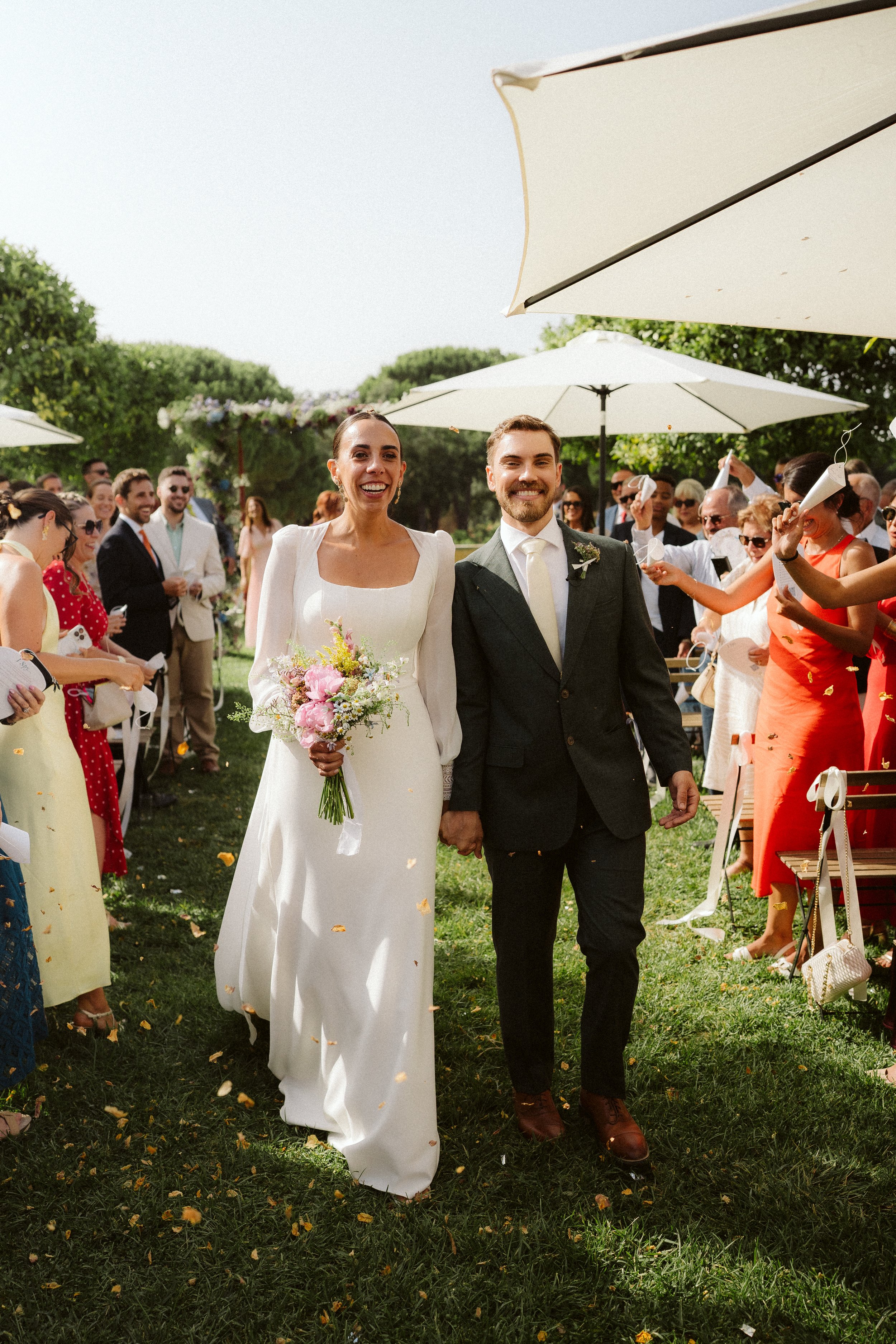 Bride and groom walking outdoors during the wedding ceremony, surrounded by guests under parasols, with trees in the background.