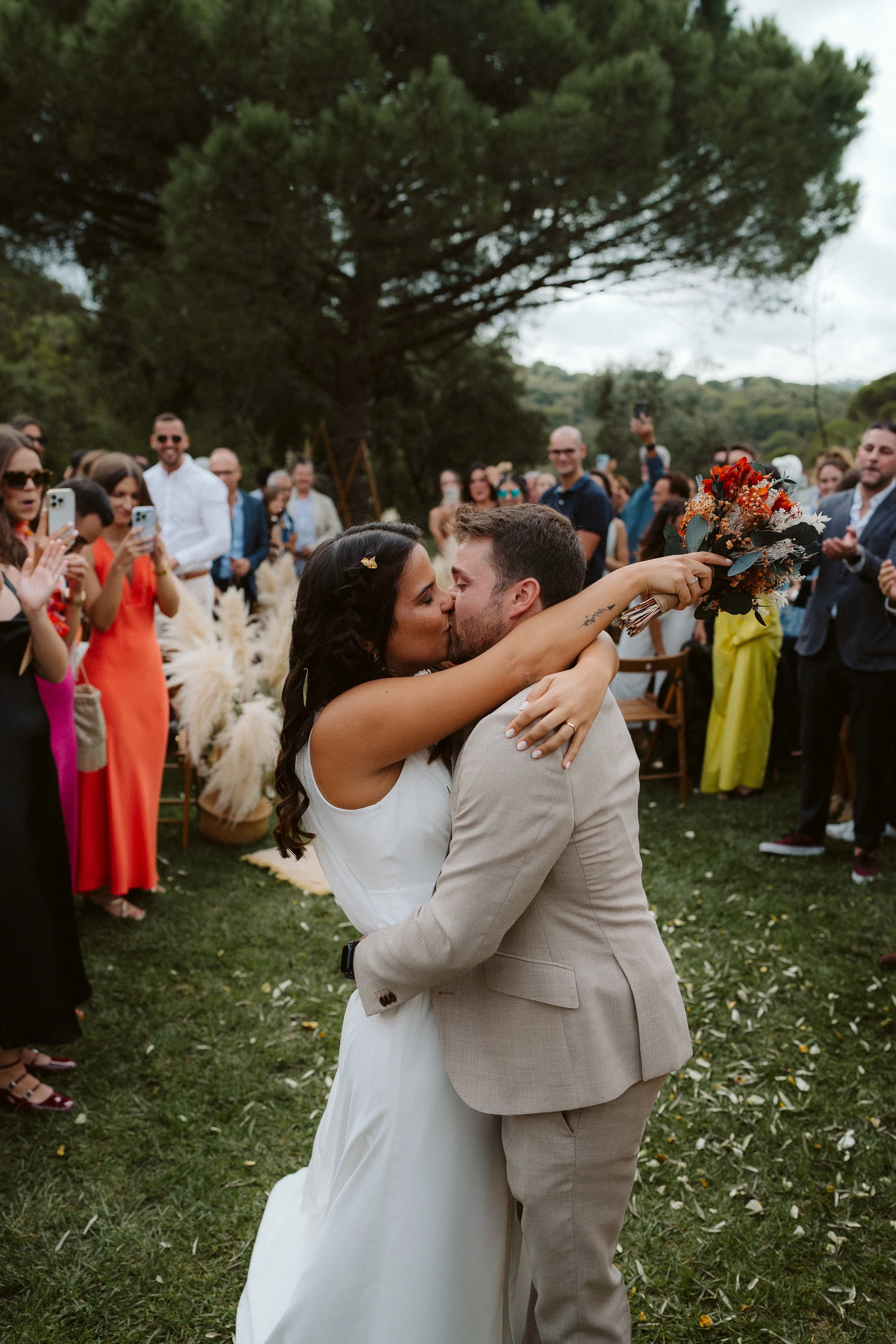 Newlywed couple kissing during an outdoor ceremony, surrounded by guests.