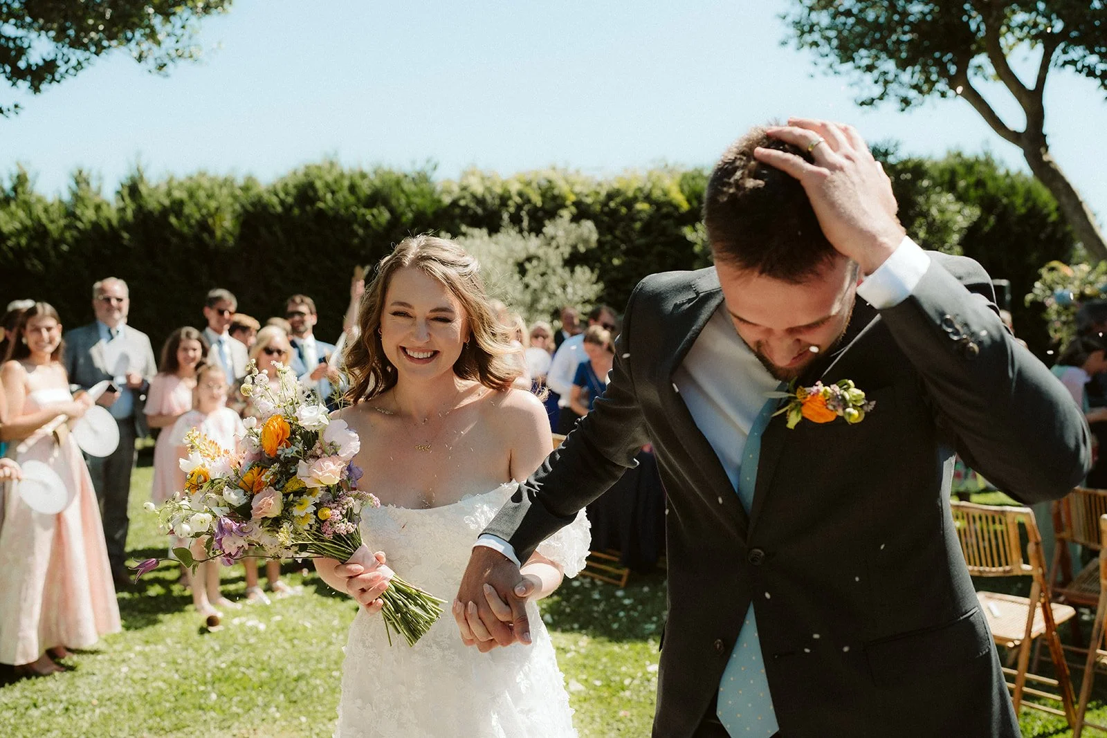 Bride and groom smiling as they say goodbye after an outdoor wedding, with happy guests in the background.