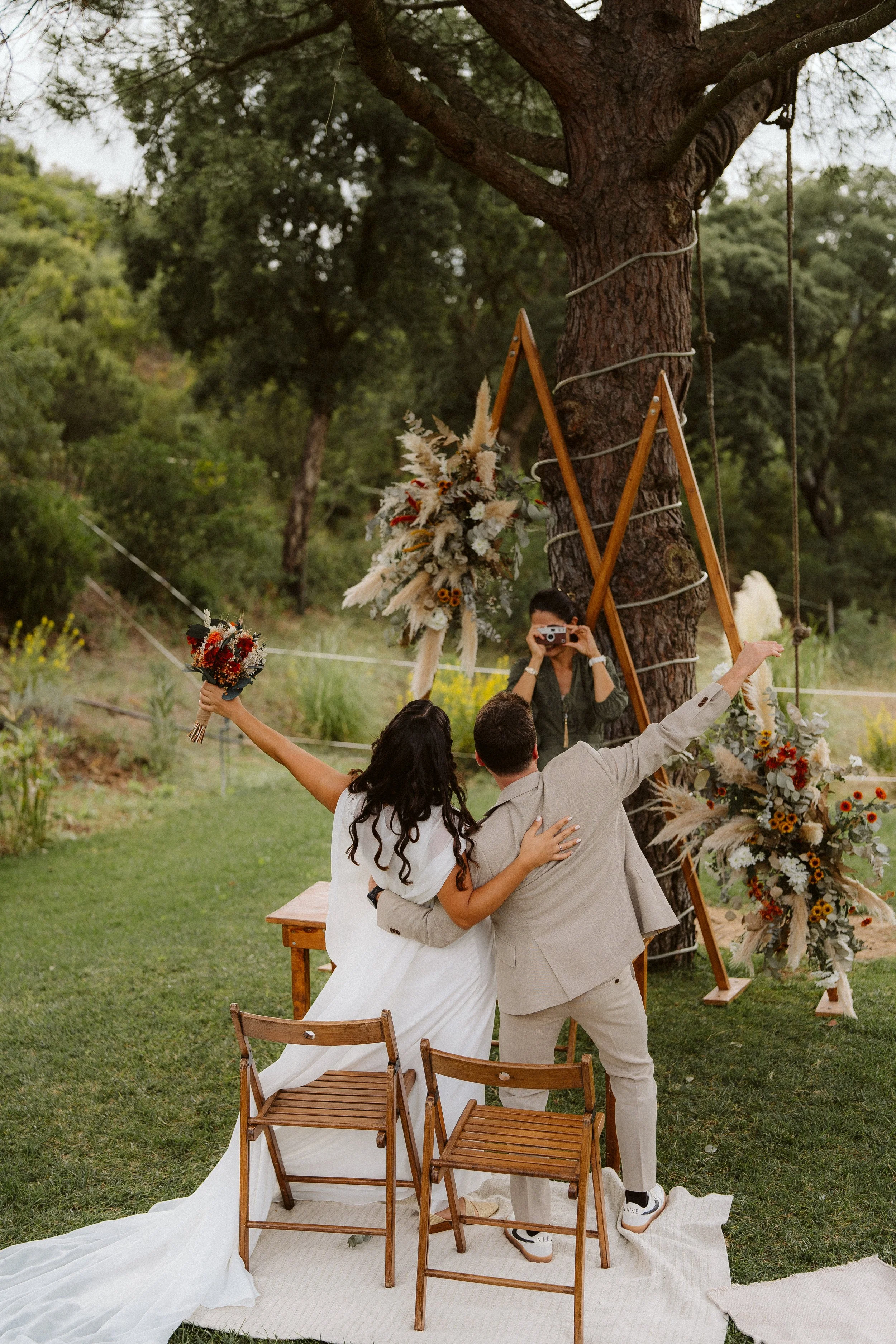 Wedding couple celebrating in nature, with floral decorations and a wooden structure, while someone takes a photo.