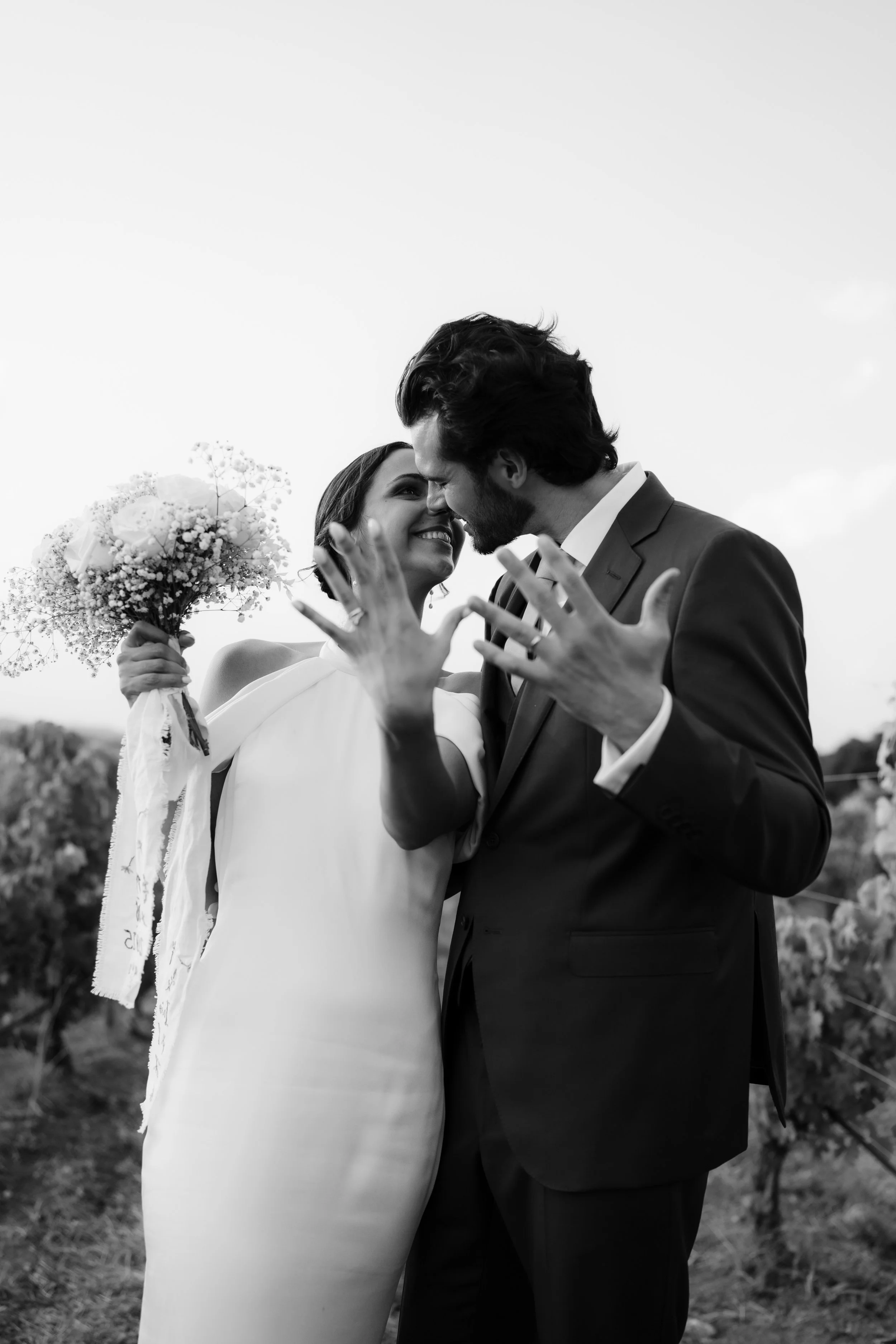 Smiling bride and groom, faces nearly touching, in a romantic outdoor setting, she holding a bouquet and wearing a white dress, he in a dark suit, in a vineyard.