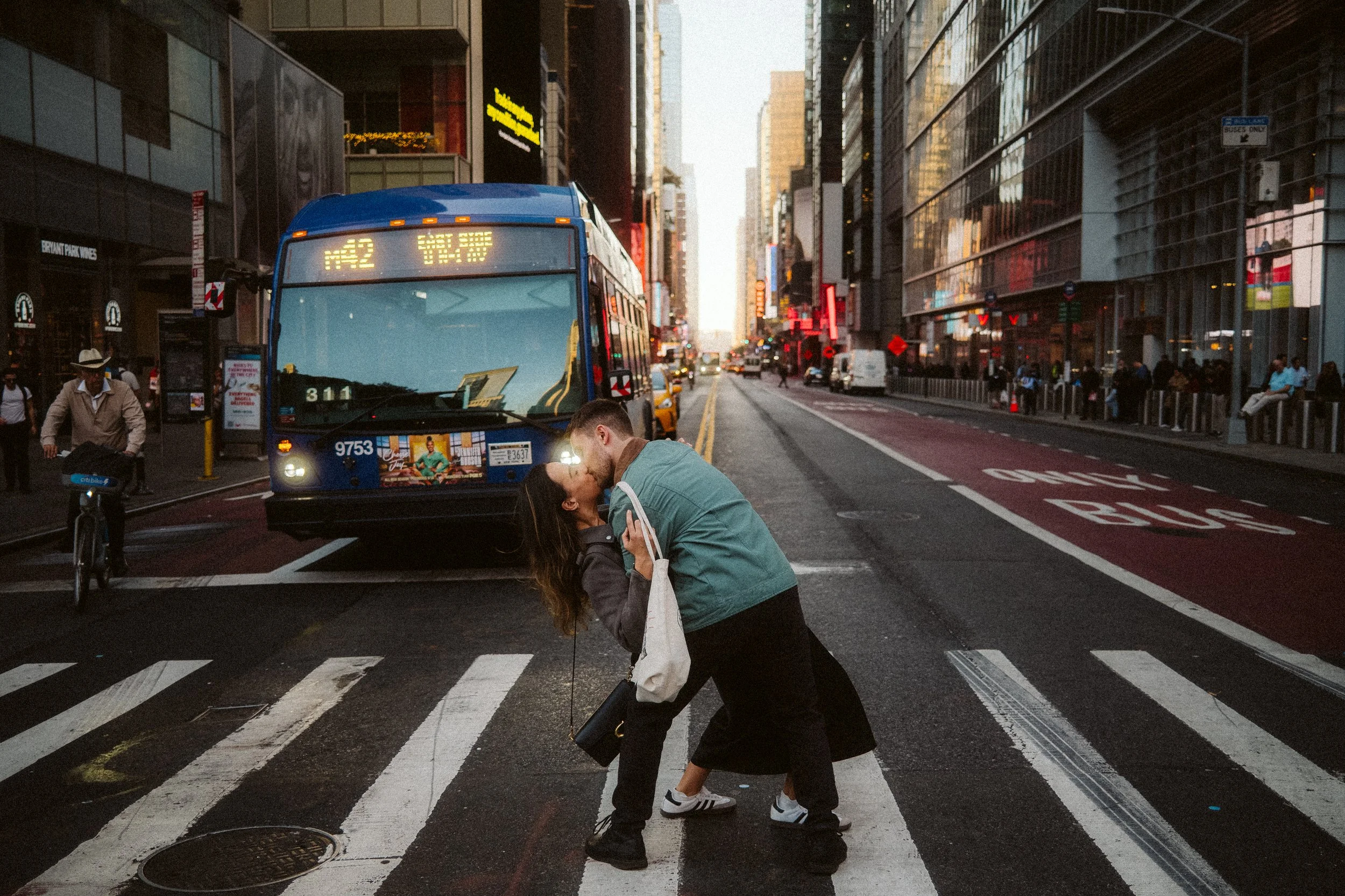 Couple kissing on a crosswalk in a busy city street, with buses and pedestrians in the background.