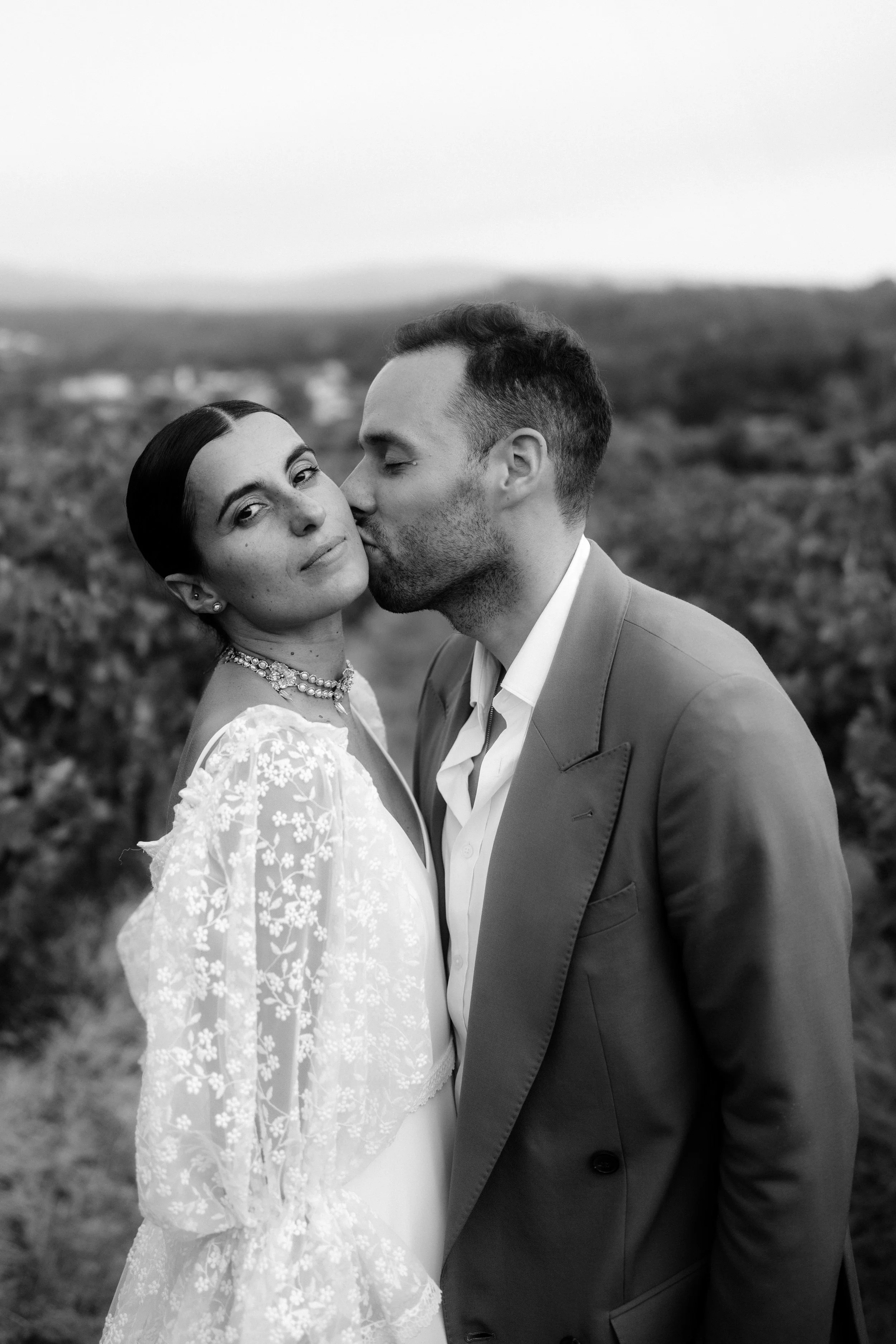 Wedding couple outdoors, the woman wearing a lace-detailed dress and a pearl necklace, the man in a suit, with greenery in the background.