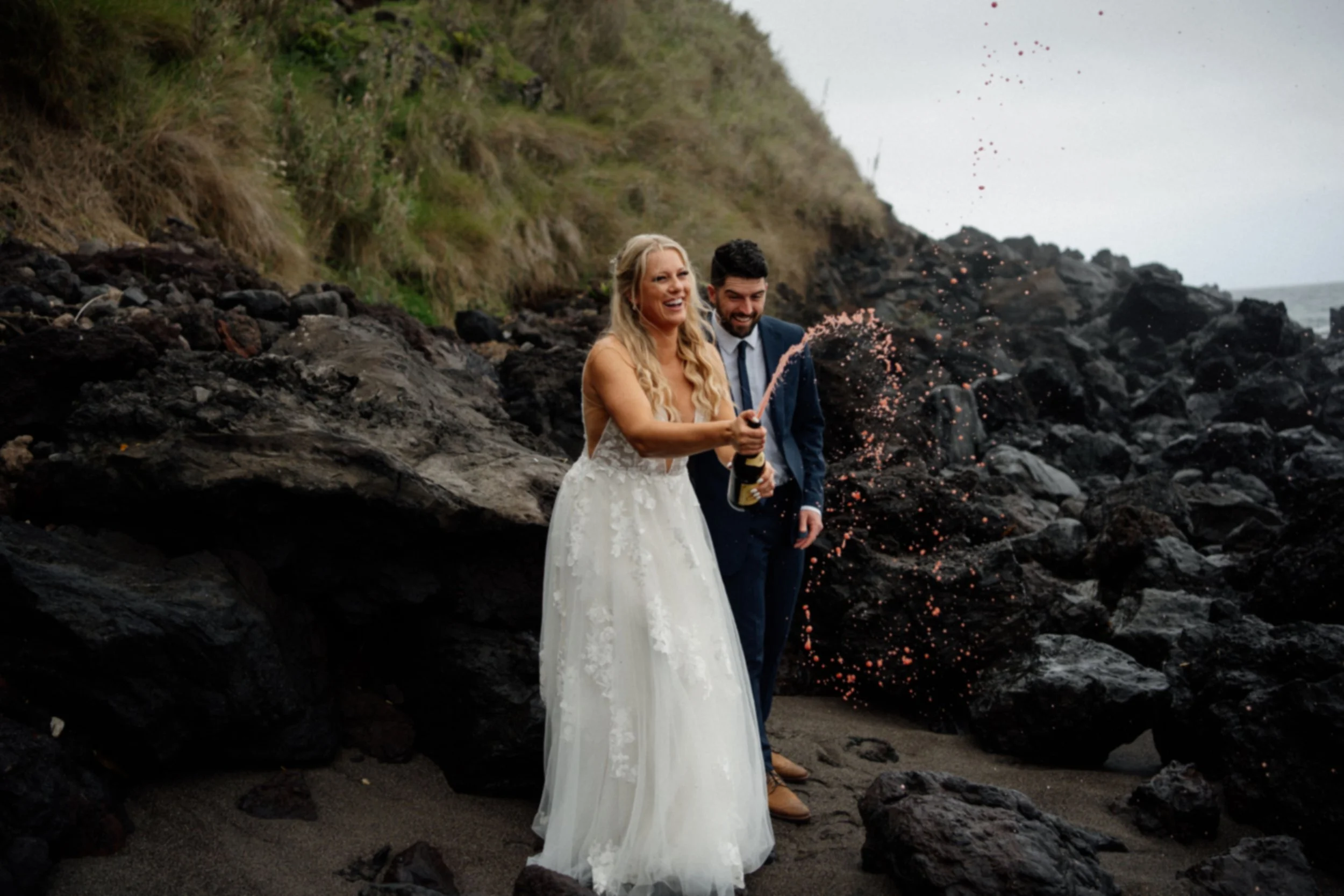Happy bride and groom on the beach, celebrating with champagne, with rocks in the background.