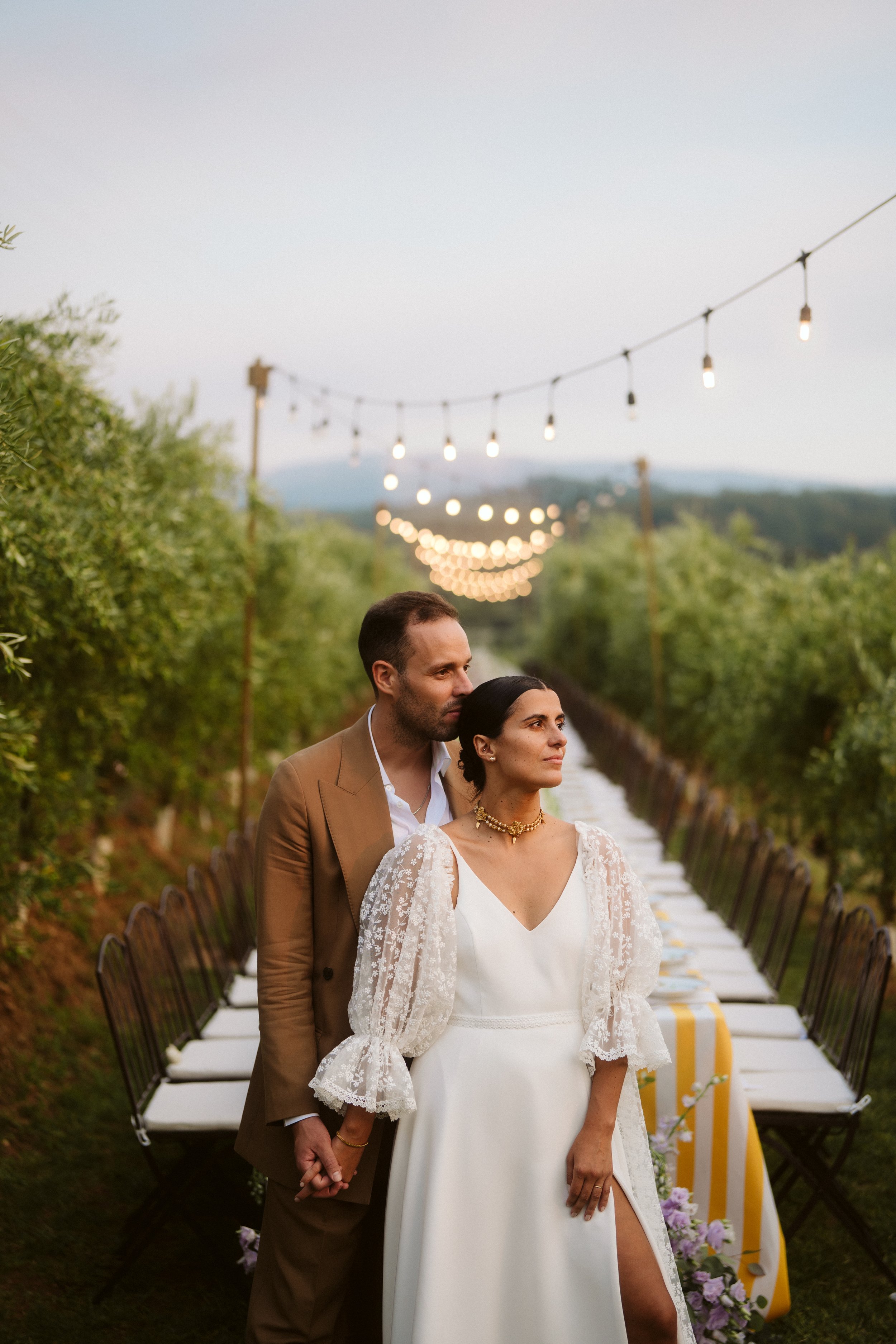 Wedding couple outdoors, holding hands, with a decorated table in the background and string lights overhead at sunset, surrounded by green trees.