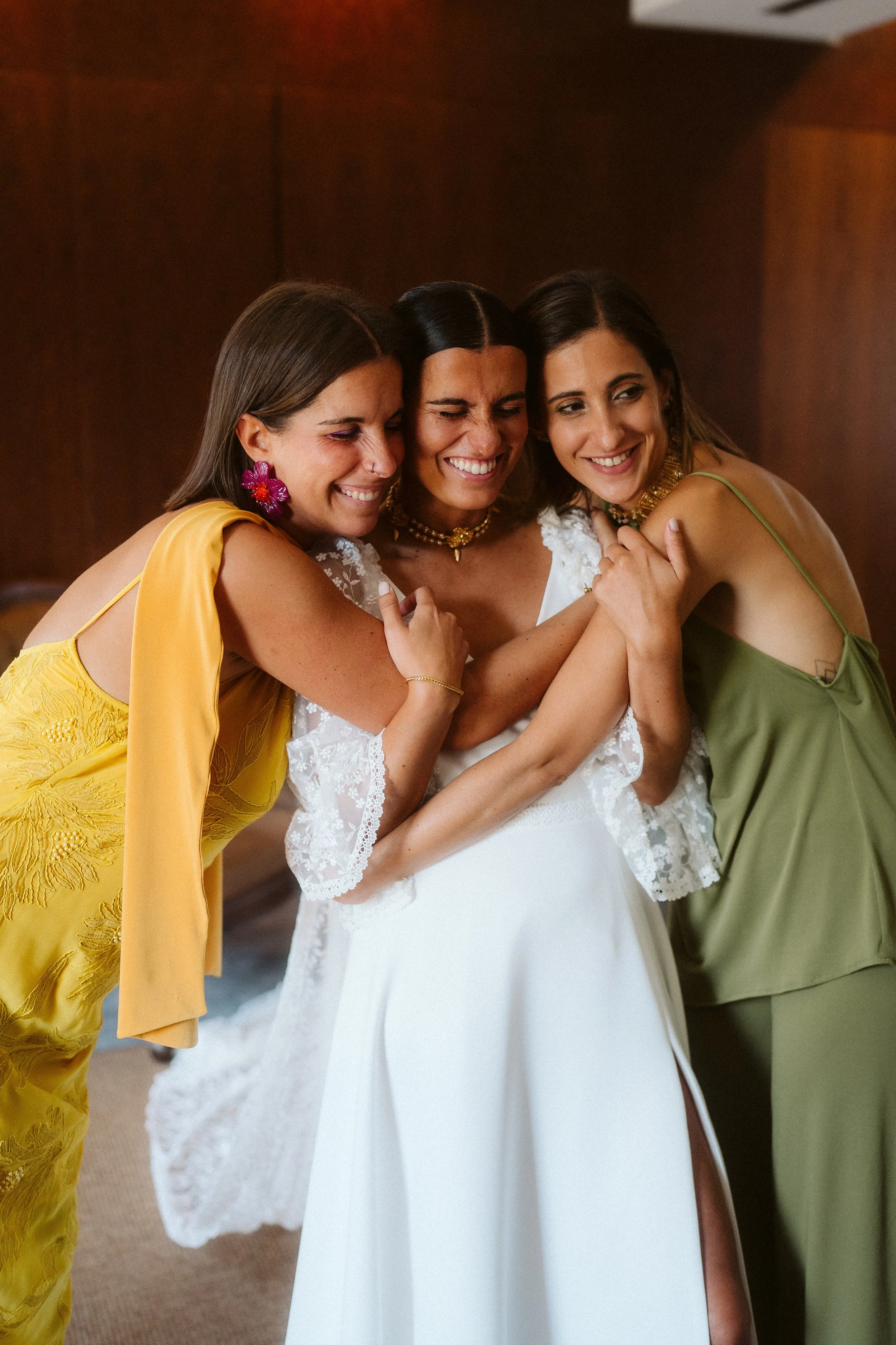 Three smiling women hugging, one of them a bride in a white lace-detailed dress, and two friends in green and yellow dresses.