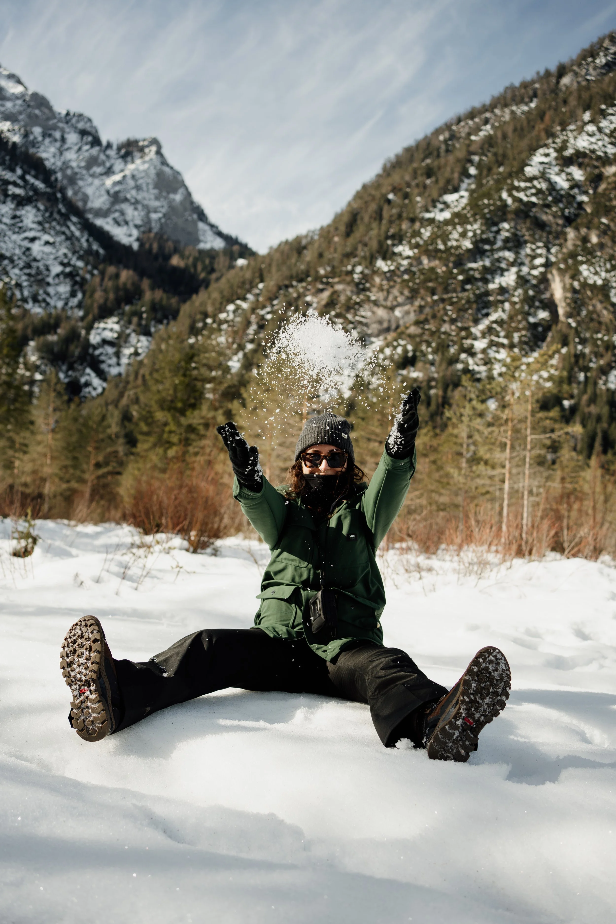 Person sitting in the snow, tossing snow into the air outdoors, with mountains and trees in the background, wearing winter clothing, sunglasses, a hat, and gloves.