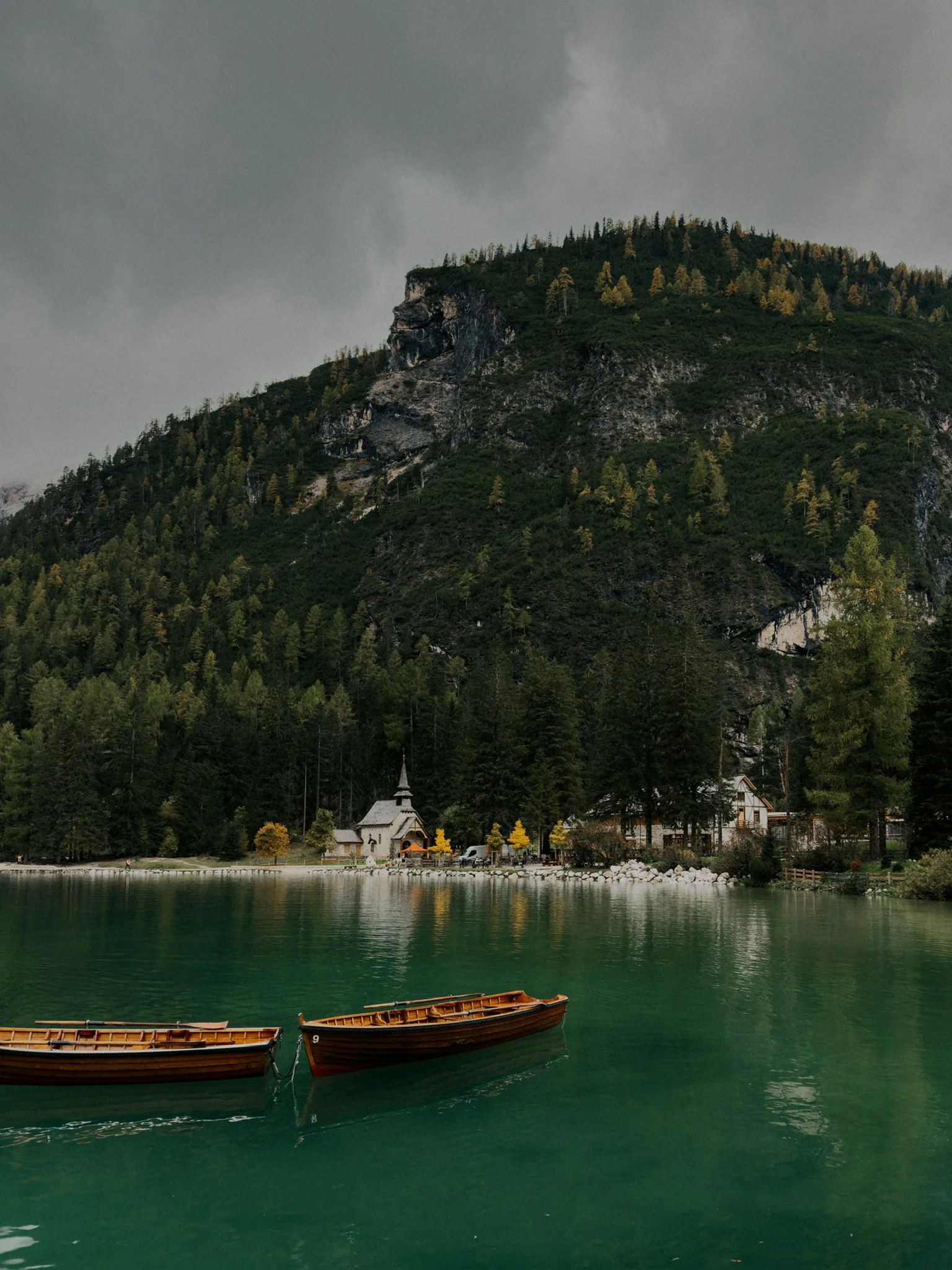 Landscape of a lake with wooden boats, surrounded by trees and a church, with a mountain in the background under a cloudy sky.