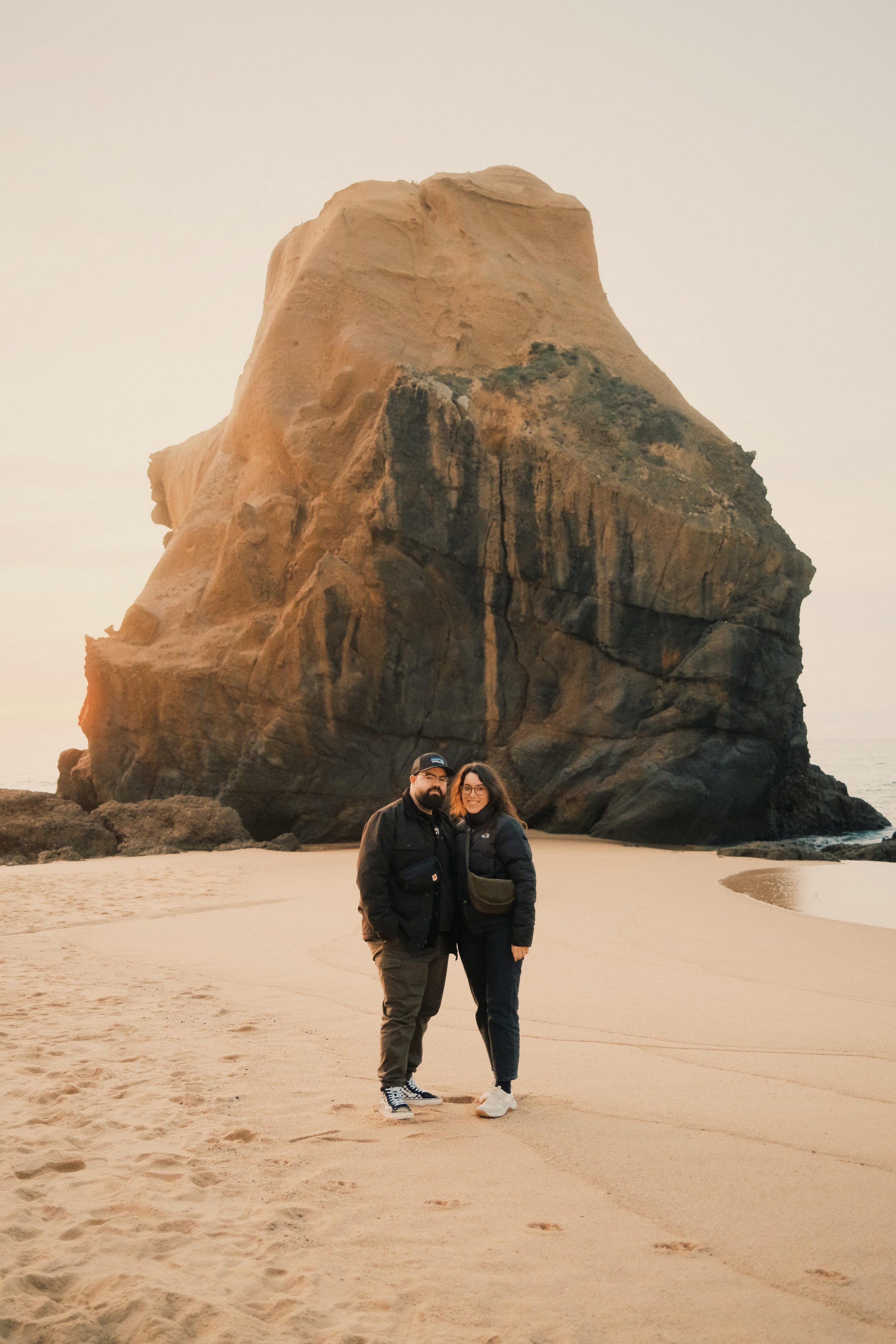 A couple posing on the beach, with a large rock formation in the background, during sunset.