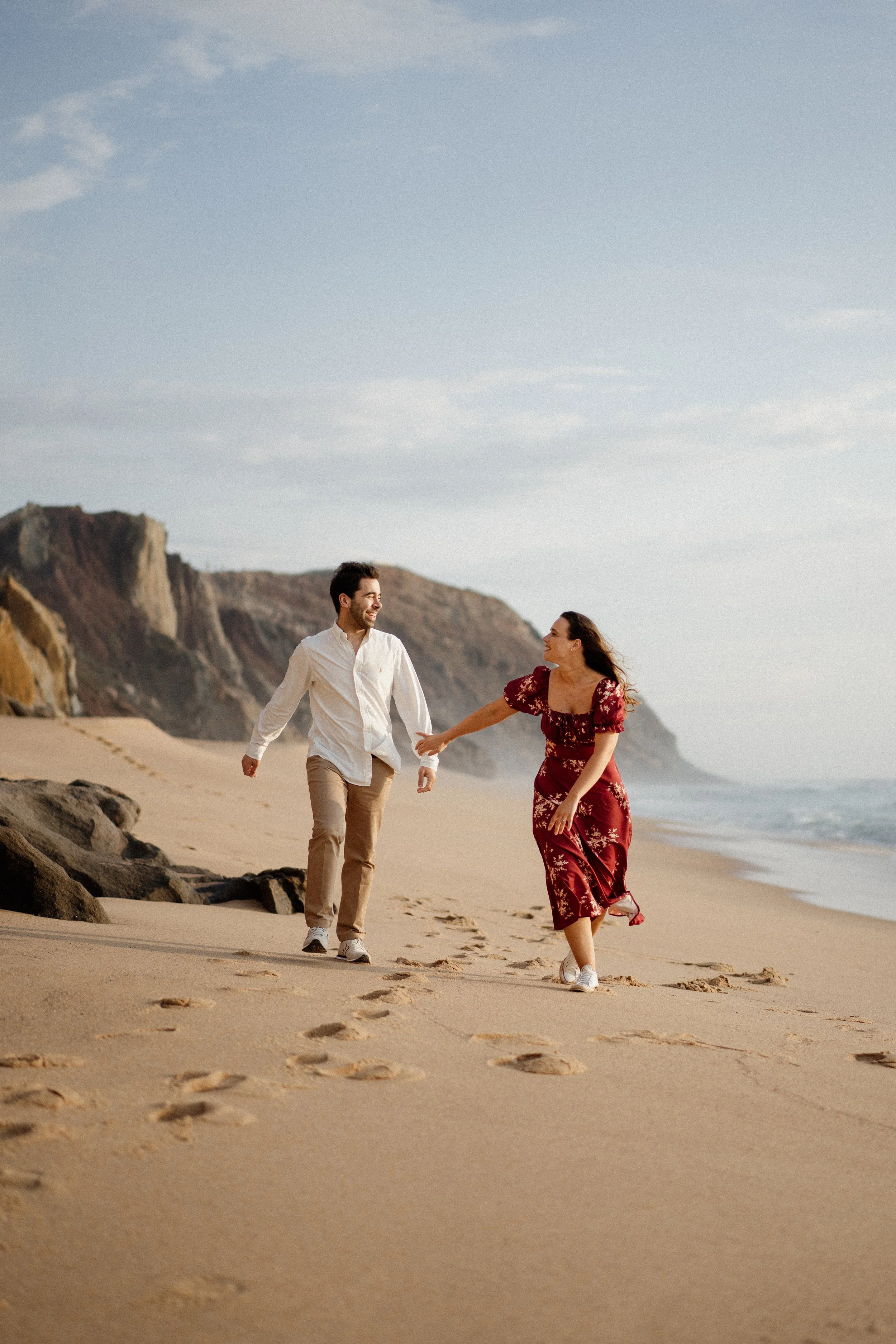 Couple walking hand in hand on the beach, with cliffs in the background, during the day.