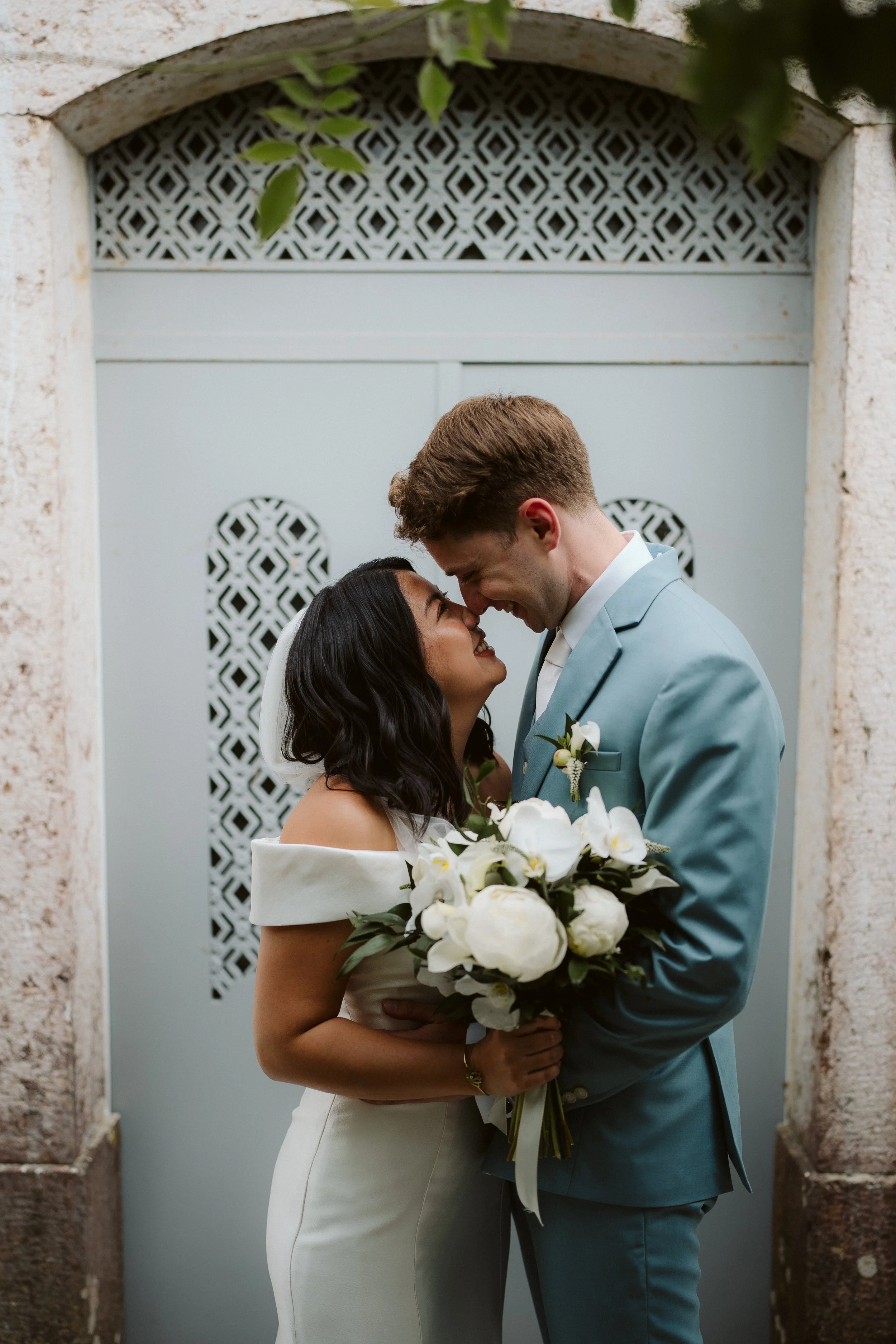 Bride and groom looking at each other and smiling, the bride holding a bouquet of white flowers, in front of a stone wall door.