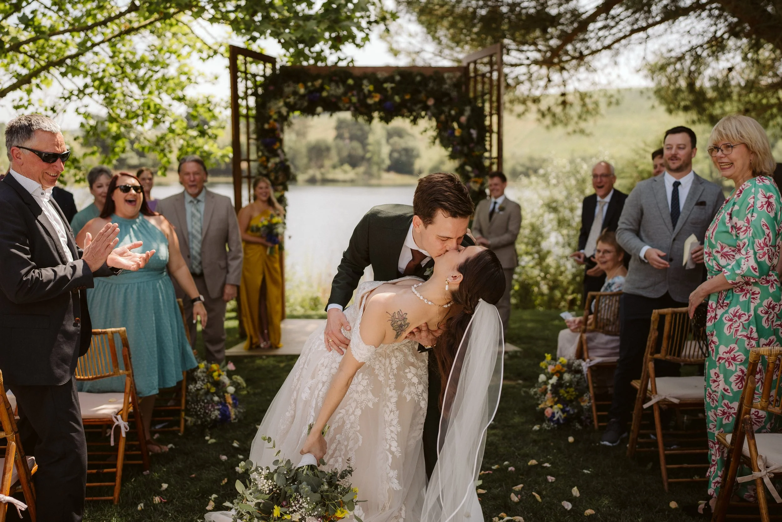 Bride and groom kissing during an outdoor wedding ceremony by a lake, with guests around them smiling and clapping.