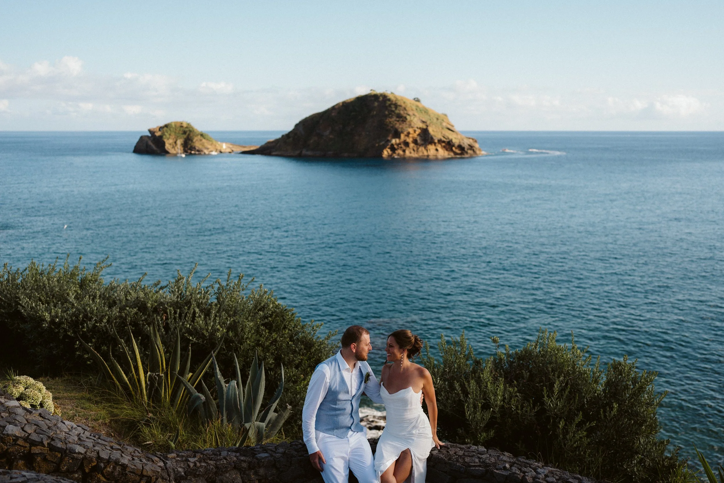 Wedding couple sitting on a stone wall by the shore, with the sea and a distant island in the background, on a clear day.
