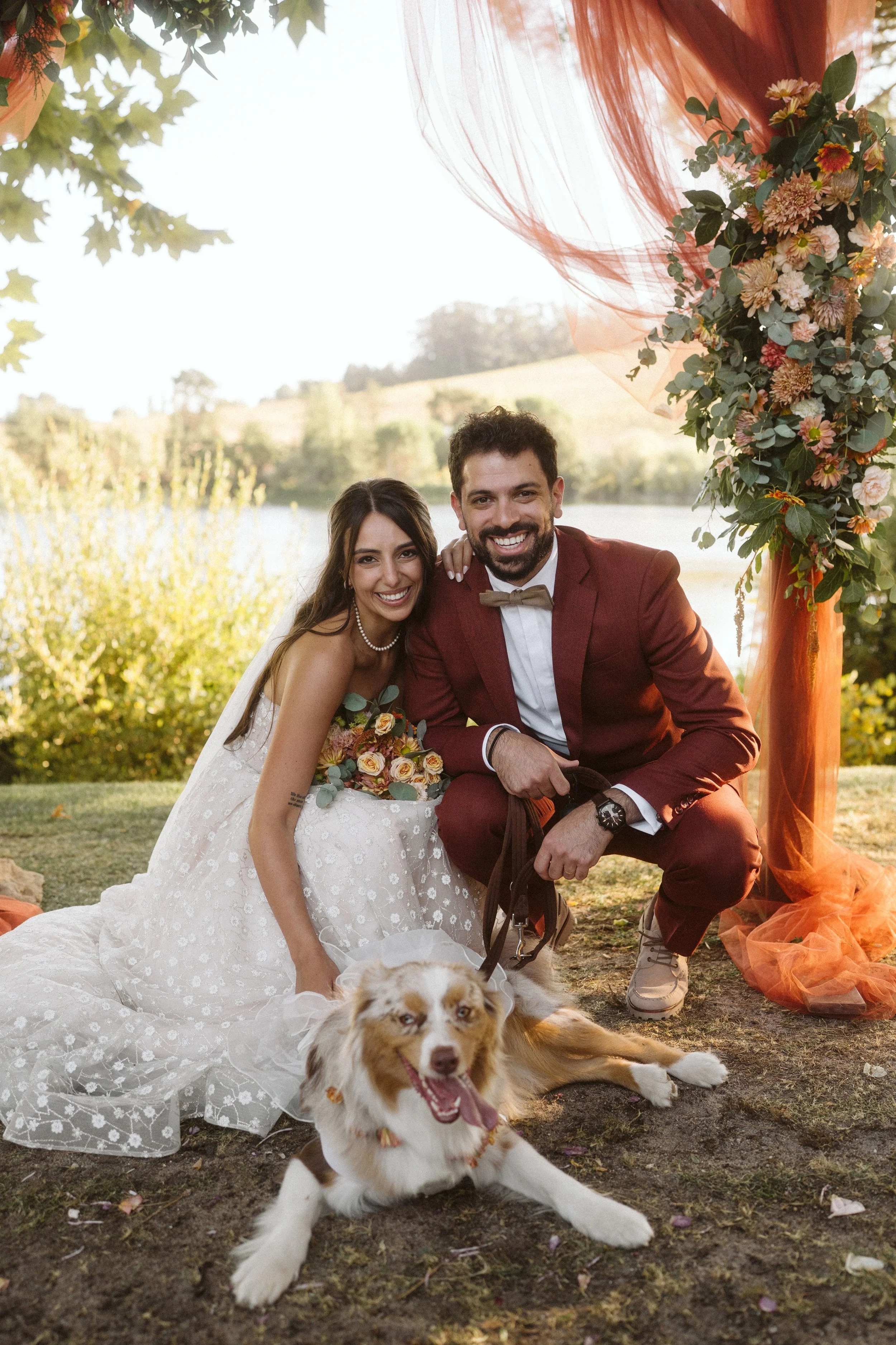 Smiling bride and groom with a dog outdoors, decorated with flowers and orange fabric, in a natural setting with a lake and trees.