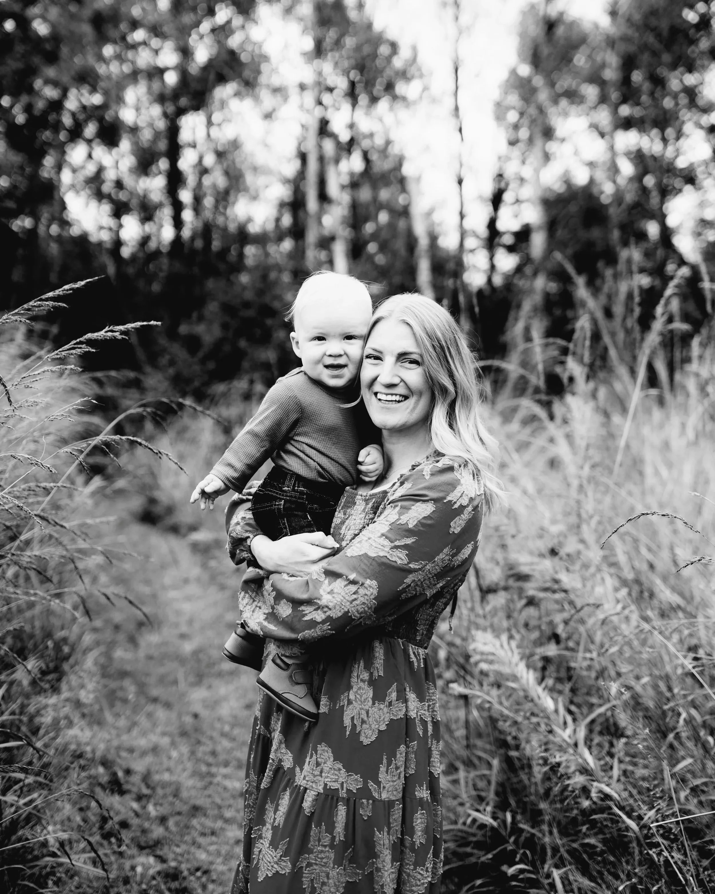 A woman holding a smiling toddler outdoors on a narrow path surrounded by tall grass and trees.