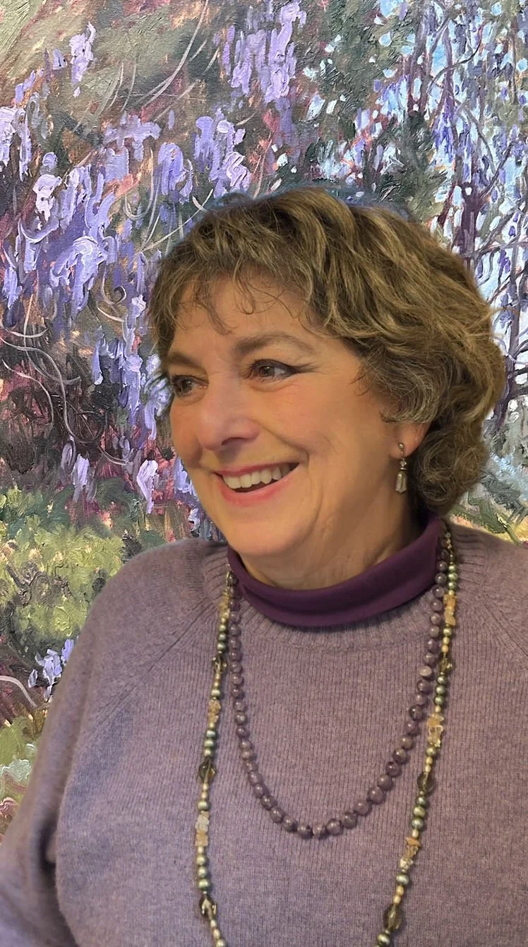A woman with short brown hair smiling, wearing a purple sweater, purple turtleneck, and layered pearl necklaces, standing in front of a colorful abstract painting with purple and green hues.