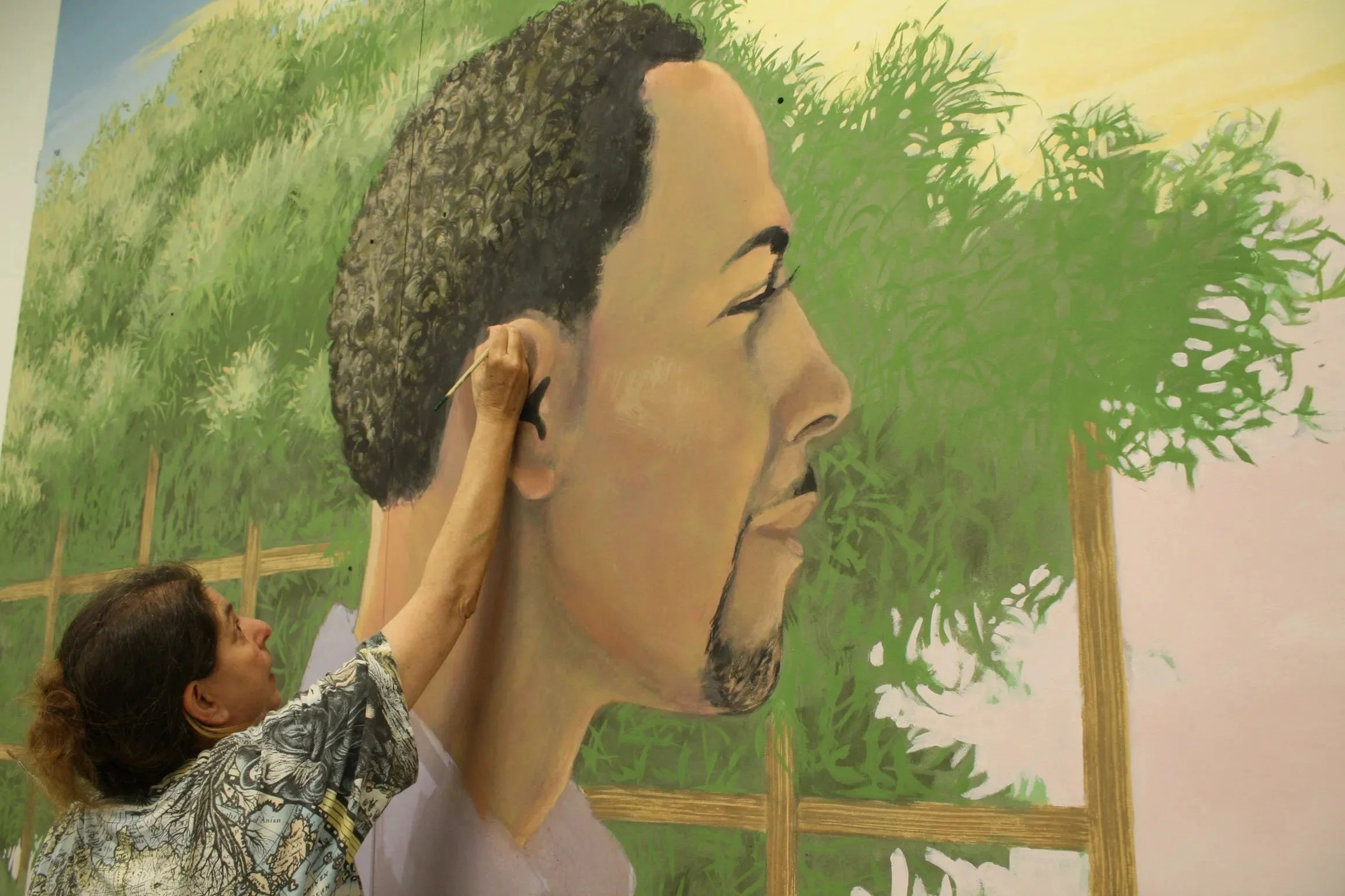 A woman painting a large mural of a man's side profile with green foliage and trees in the background.
