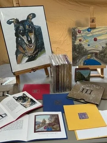 A table with a dog painting on a small easel, some open books, notebooks, a small painting on a stand, and several sealed books or DVDs.