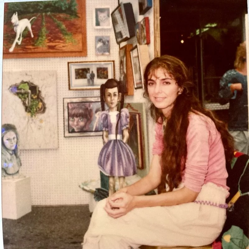 A young woman with long brown hair and a pink shirt sitting in an art gallery, surrounded by various paintings and sculptures, including a Victorian-style doll dressed in a purple gown.