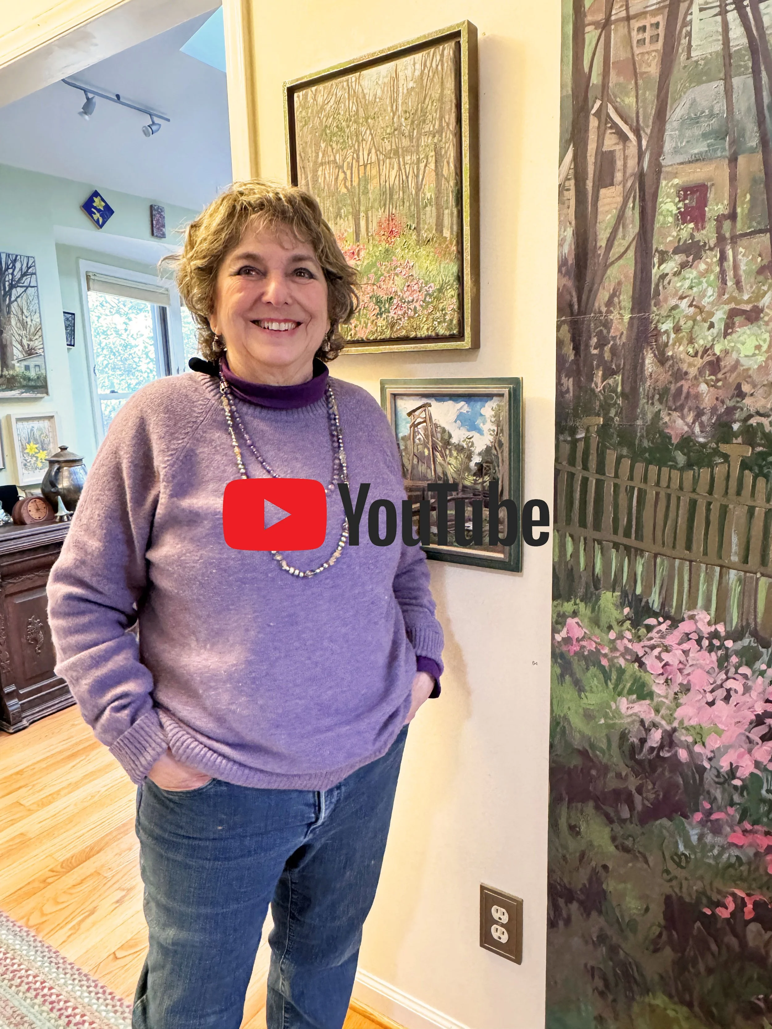 Smiling woman in purple sweater and blue jeans standing in an art-filled room, with paintings on the wall and a colorful tree painting on the right.