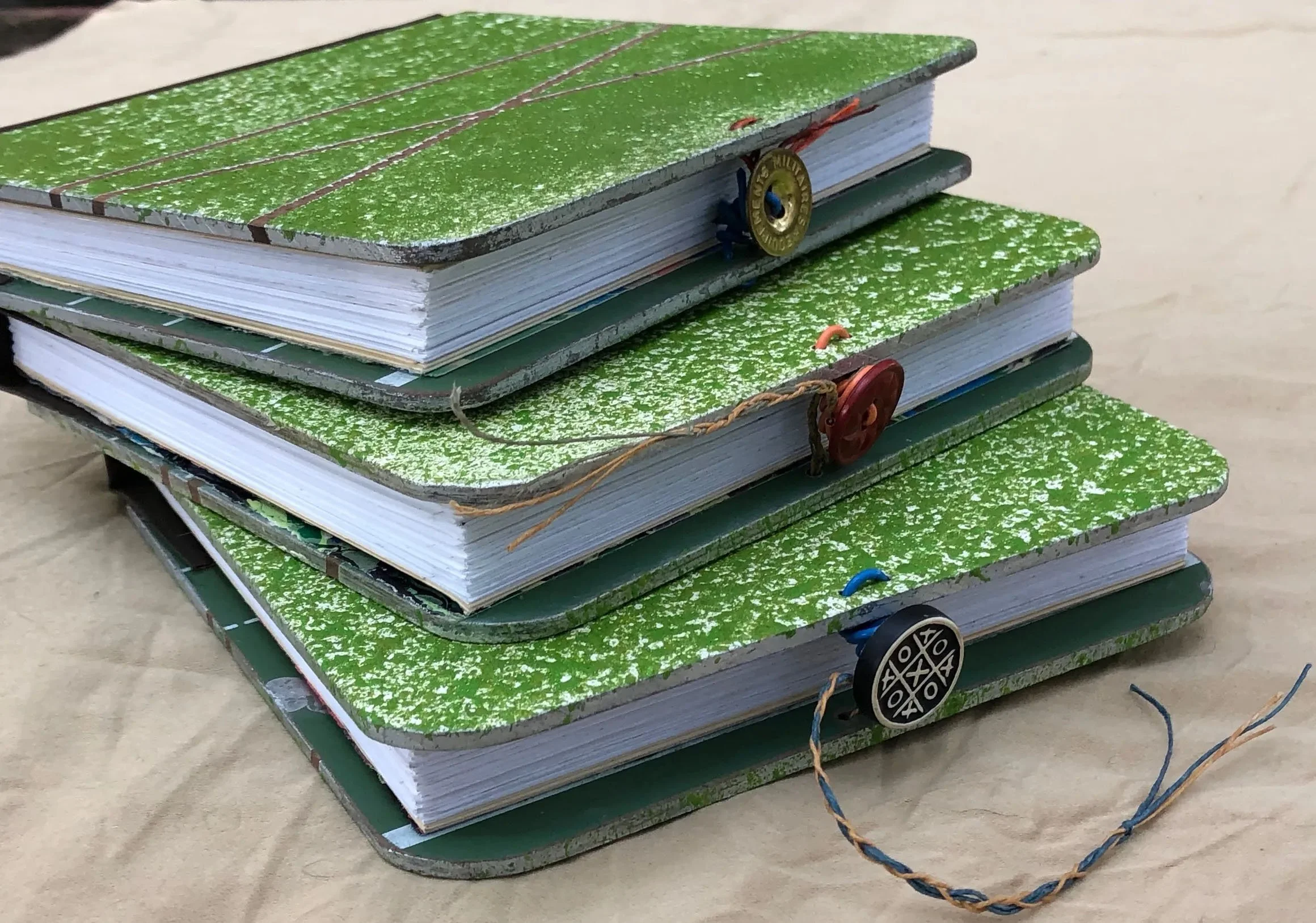 Stack of four green notebooks with speckled covers, decorated with various colored buttons and strings, placed on a wooden surface.