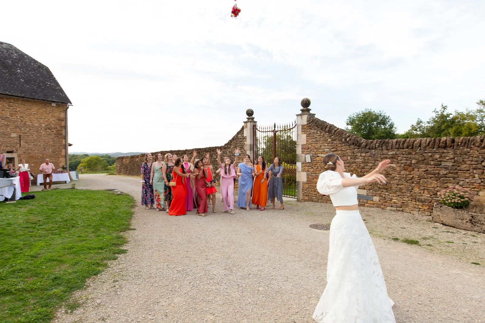 Un groupe de femmes habillées en robes colorées participe à une activité de mariage en extérieur, avec des décorations et un mur en pierre en arrière-plan.